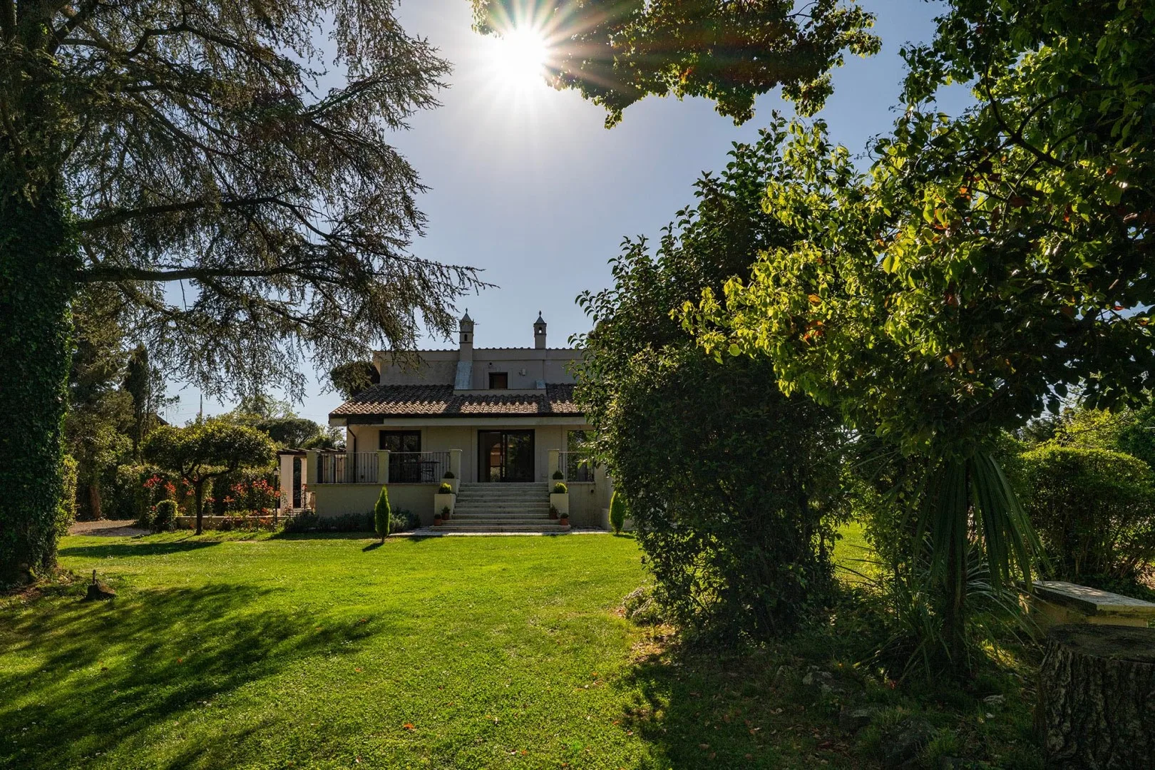A house stands beyond a sunlit lawn, surrounded by mature trees and greenery.