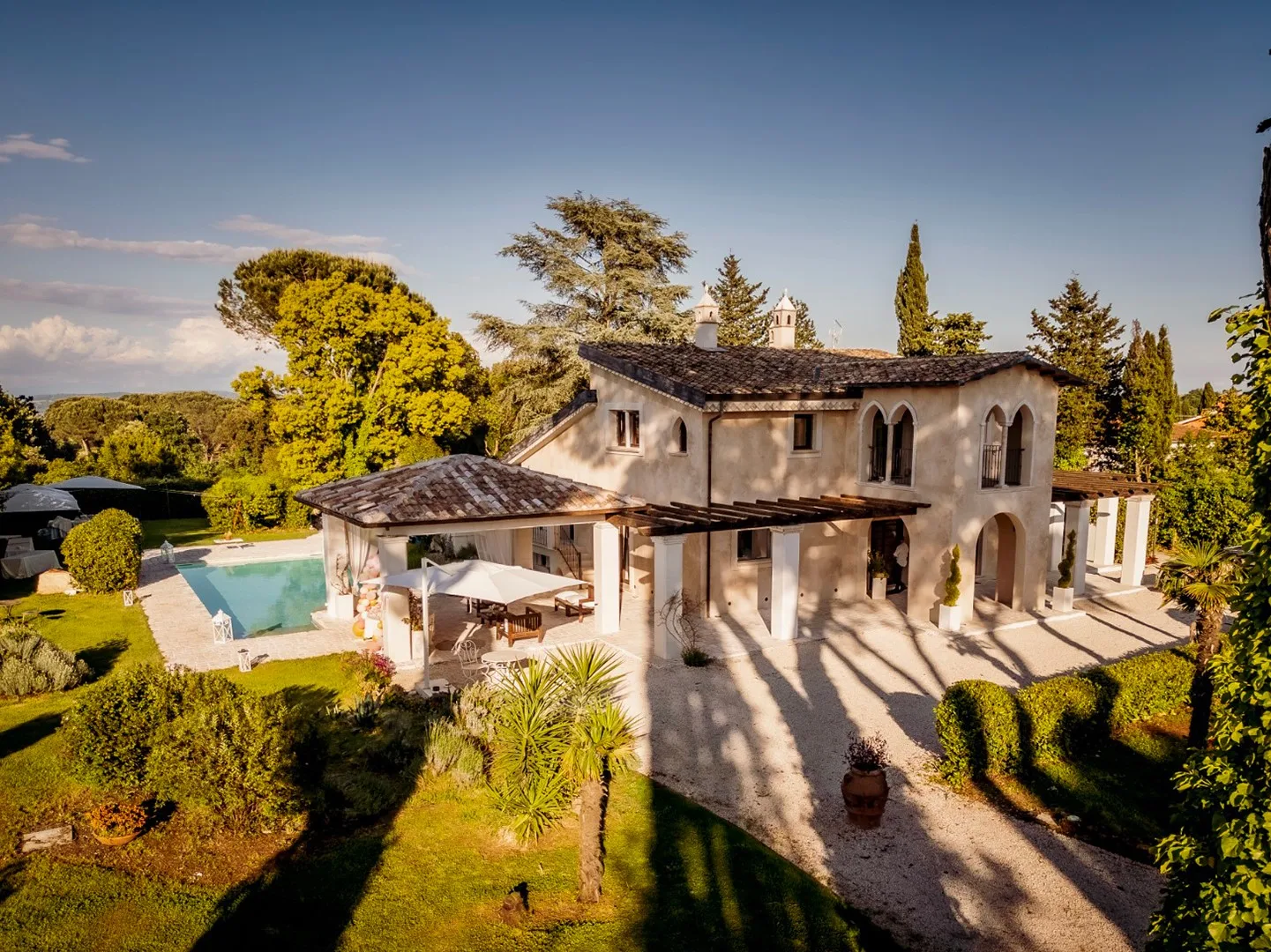 A large house sits beside a swimming pool, surrounded by trees and landscaped grounds.