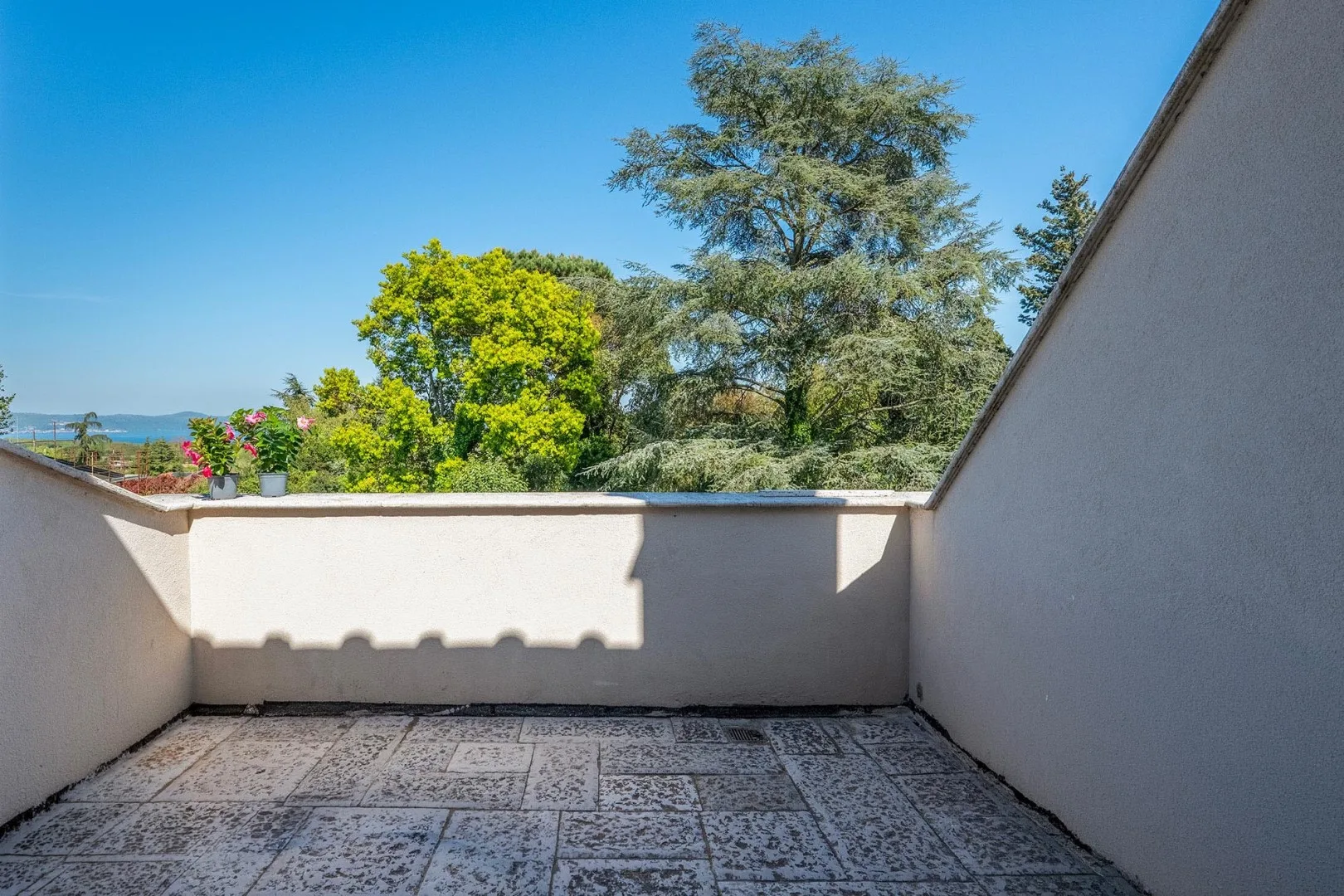 A sunlit terrace with tiled flooring and trees visible beyond the low wall.