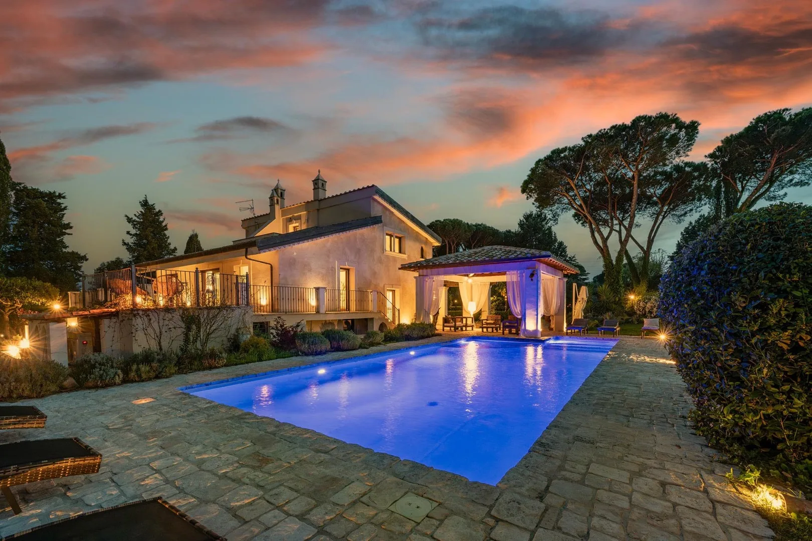 A modern house and illuminated swimming pool sit beneath a colorful sunset sky.
