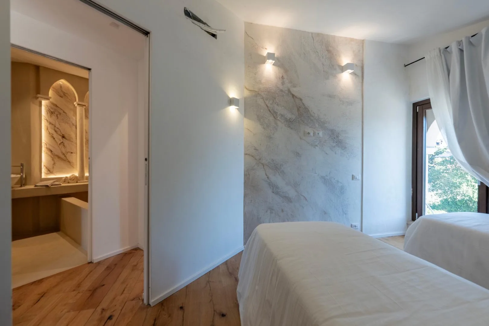 A bedroom with two beds beside a marble accent wall and an open bathroom doorway.