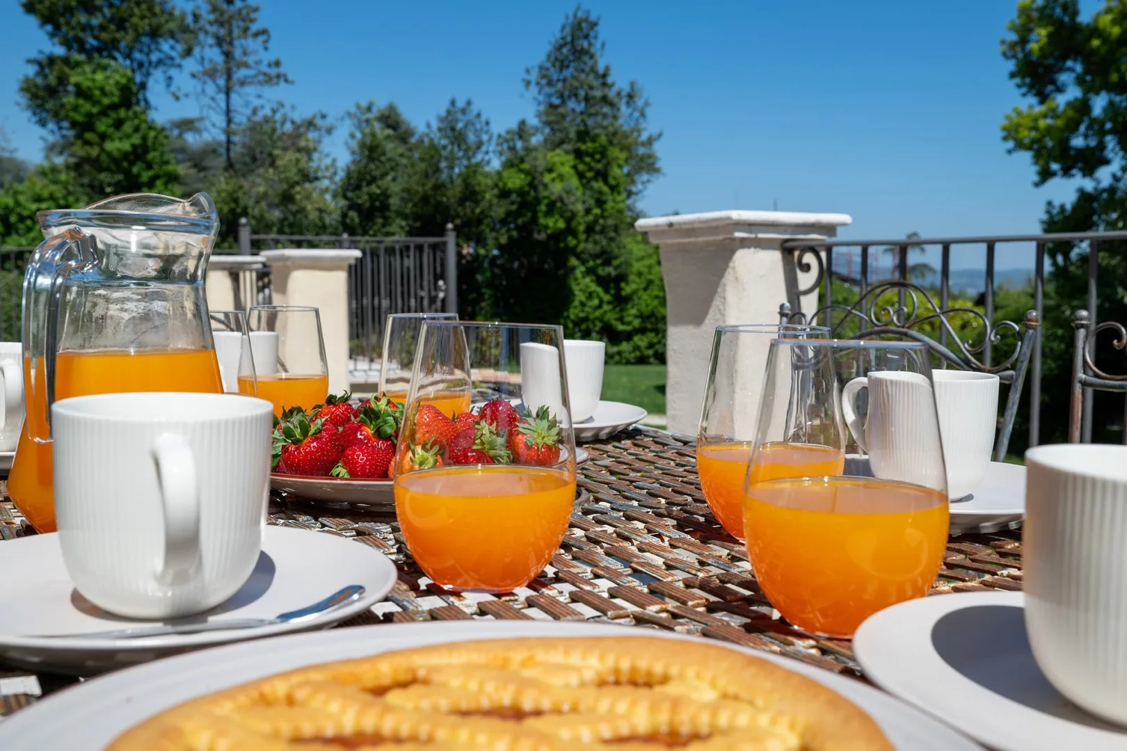 A sunny outdoor dining table is set with orange juice, strawberries, and pastries.