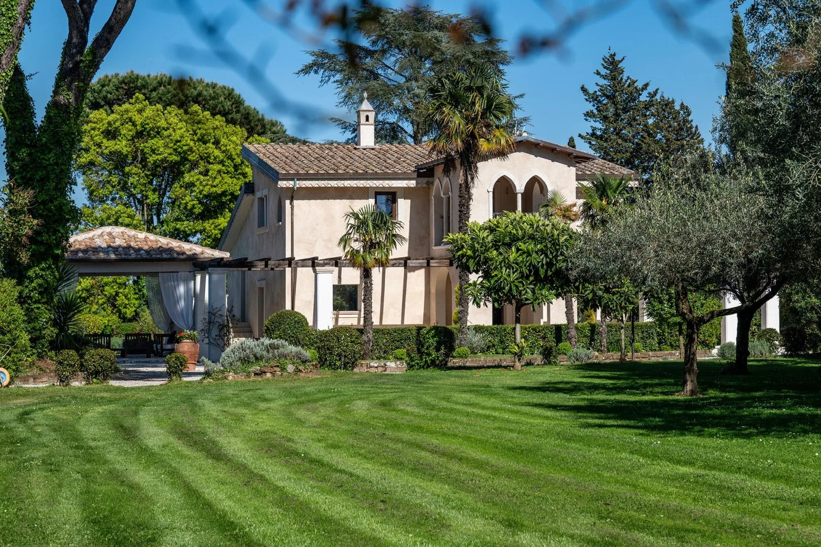 A large house stands beyond a green lawn, surrounded by trees and shrubs.