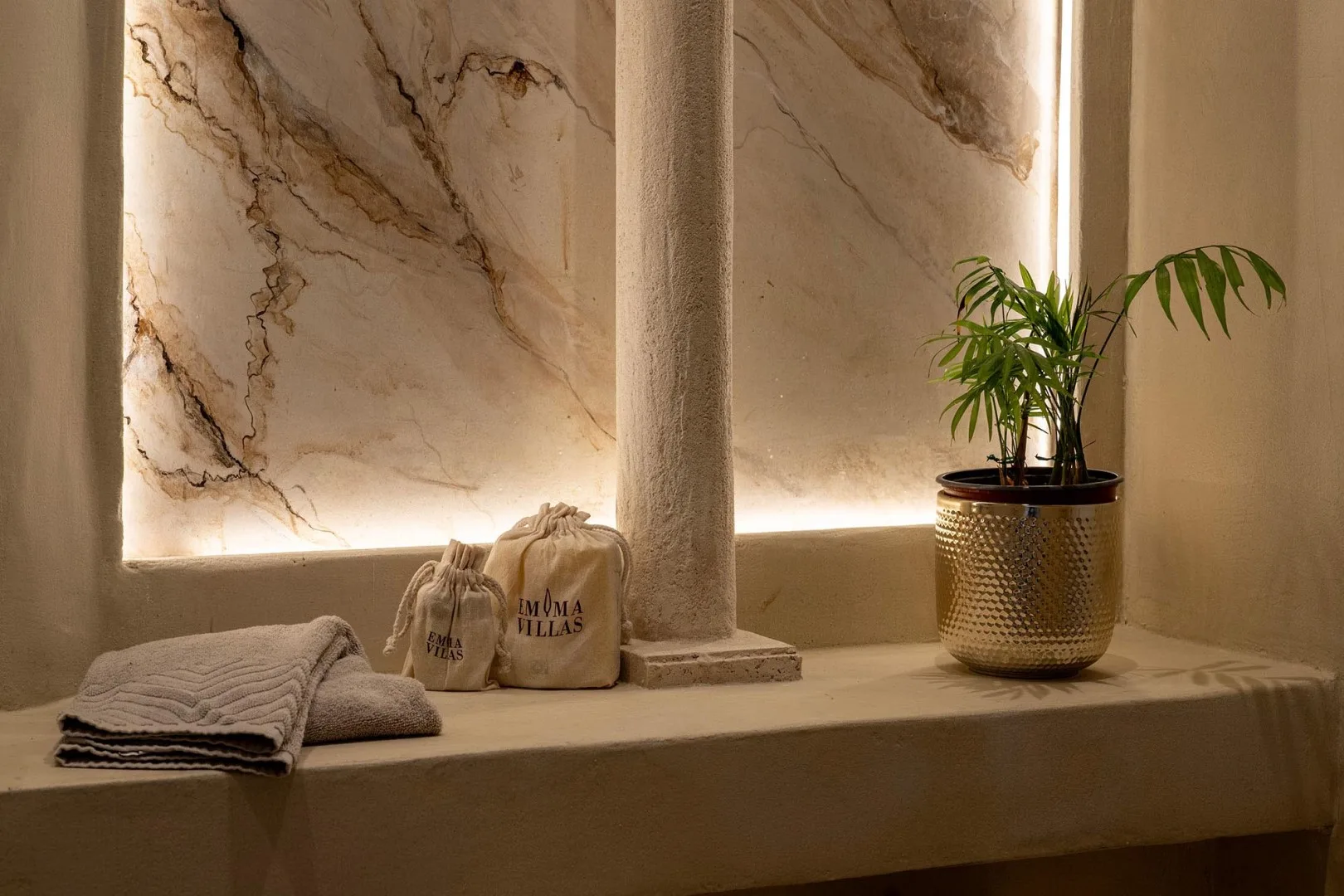 A softly lit marble wall display with folded towels, fabric bags, and a potted plant.