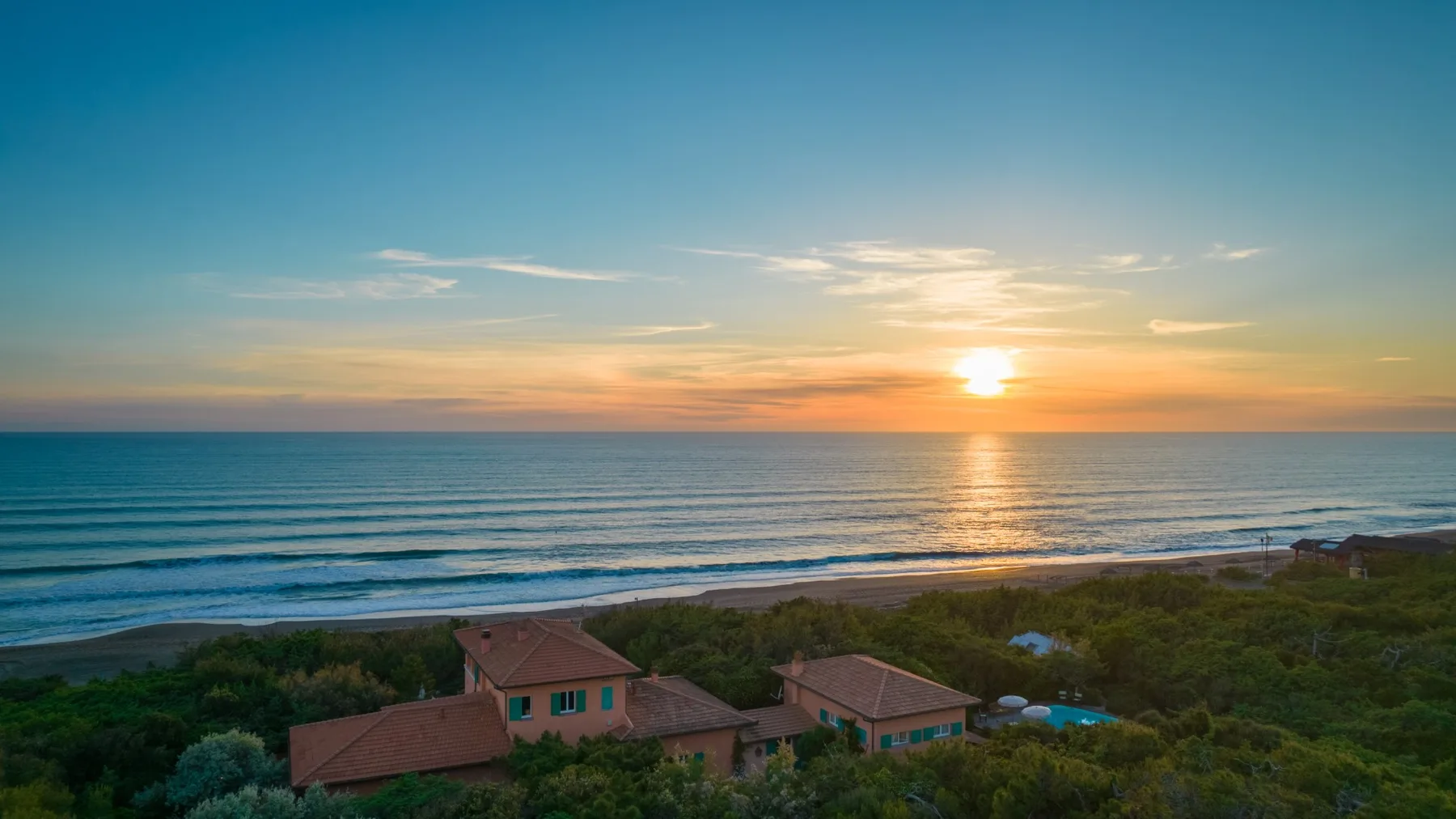 A coastal scene at sunset, with waves rolling toward houses among green vegetation.
