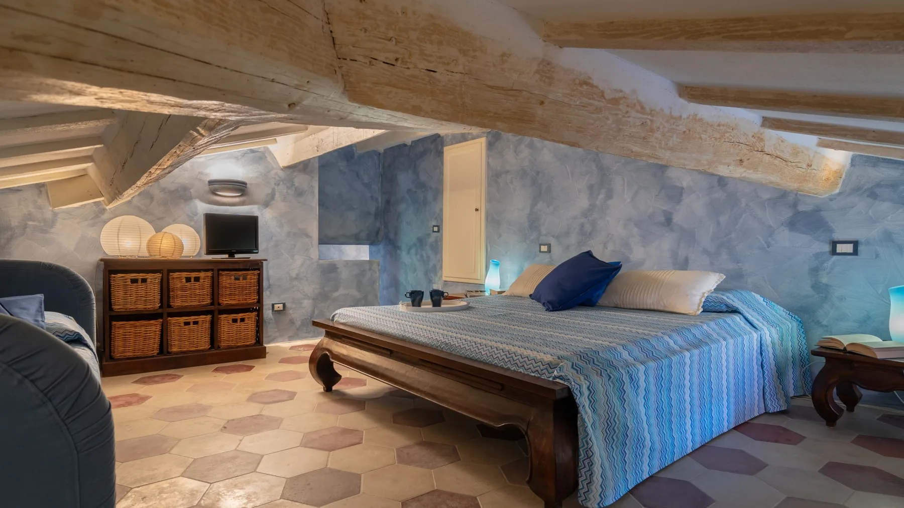 A bedroom with a bed beneath exposed wooden beams and a patterned floor.