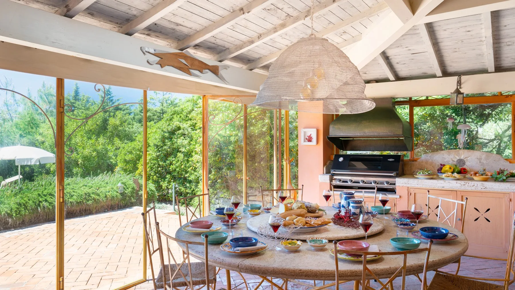 A dining area with a long table sits beside large windows overlooking greenery.