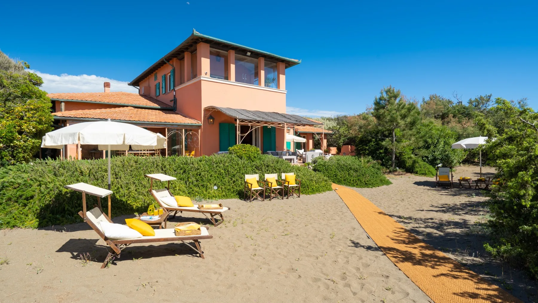 A sandy outdoor seating area beside a two-story building, with loungers and greenery.