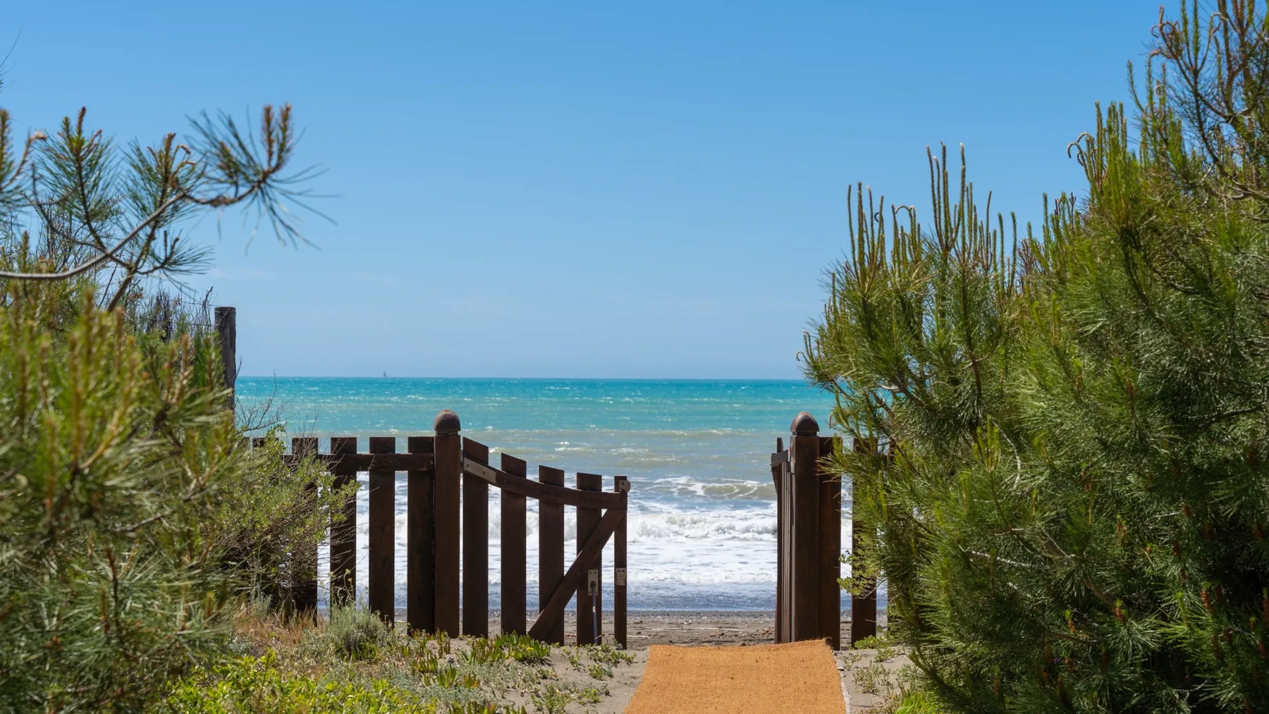 A sandy path leads through a wooden gate toward the ocean, bordered by coastal plants.