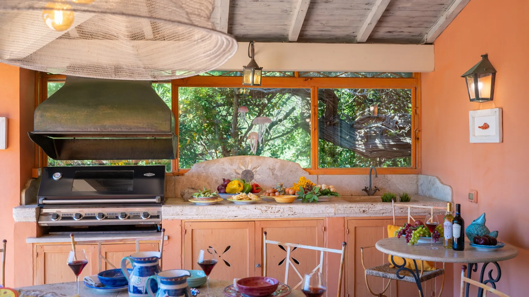 An outdoor kitchen with a grill, counter, and windows overlooking greenery.