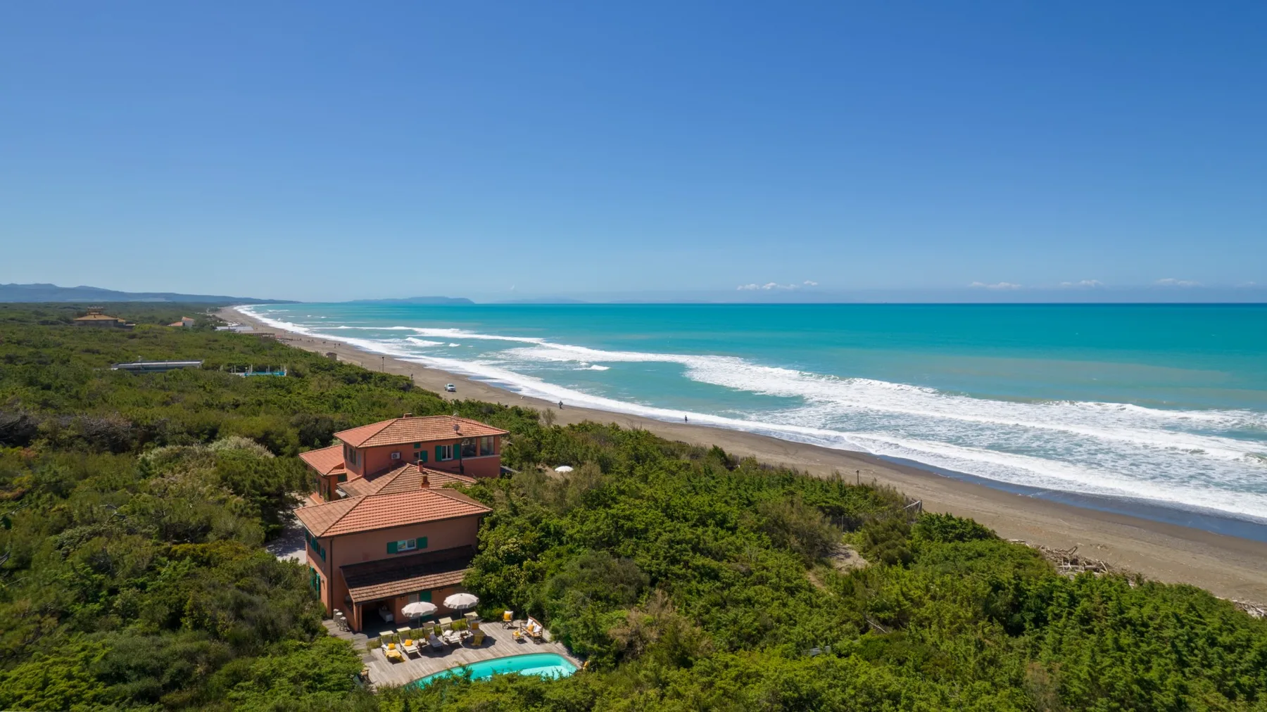 A coastal house sits among green vegetation beside a long sandy beach and turquoise ocean.