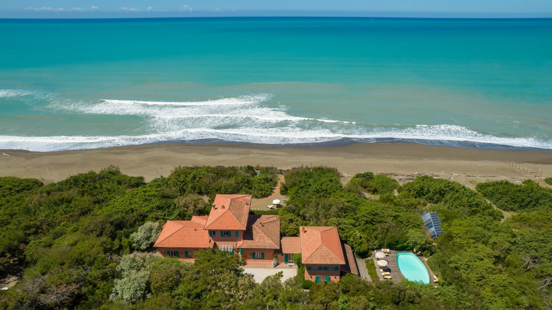 Aerial view of a seaside house amid greenery, with a pool near the beach.