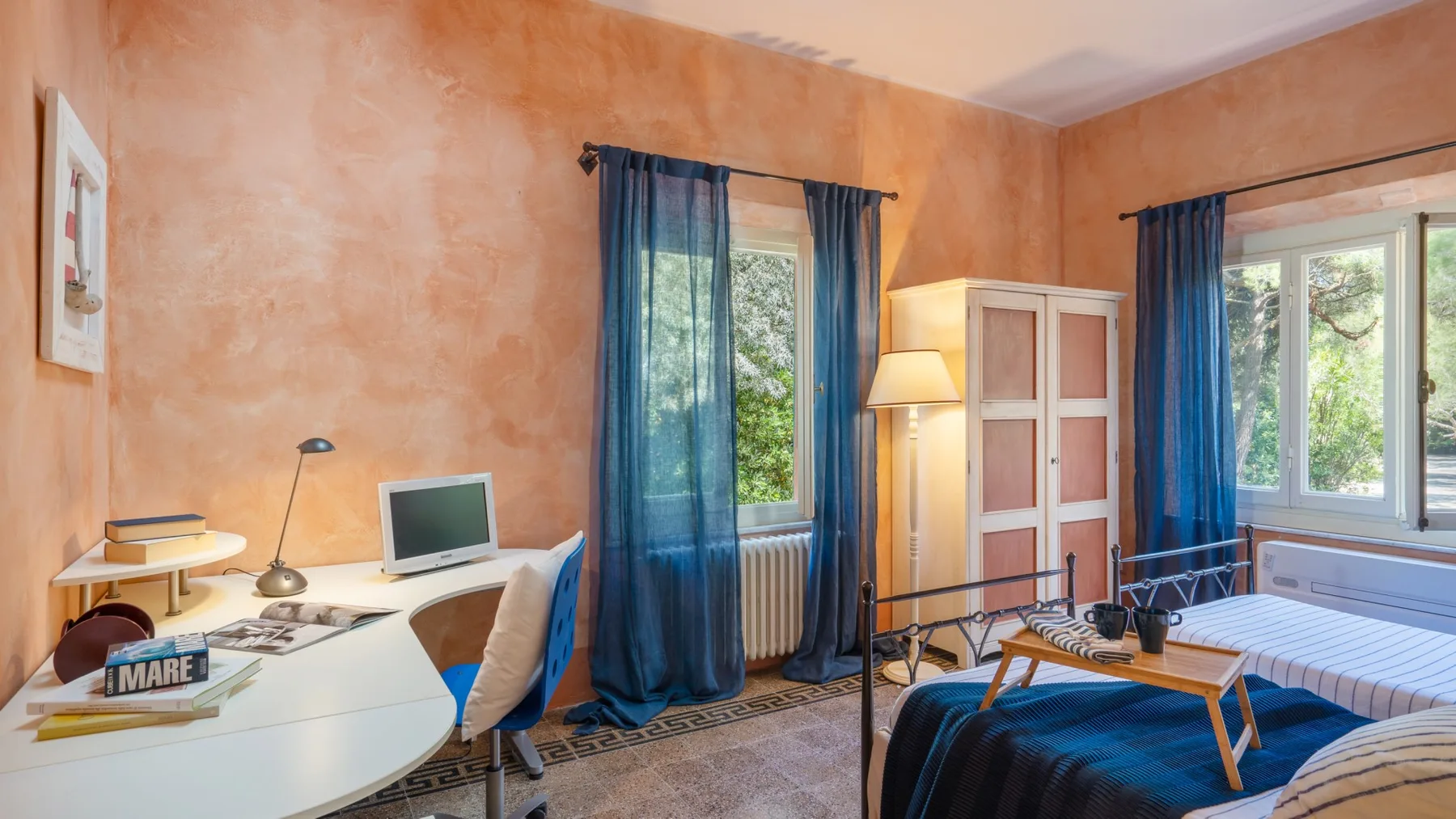 A bedroom with a desk, computer, and blue curtains beside the bed.