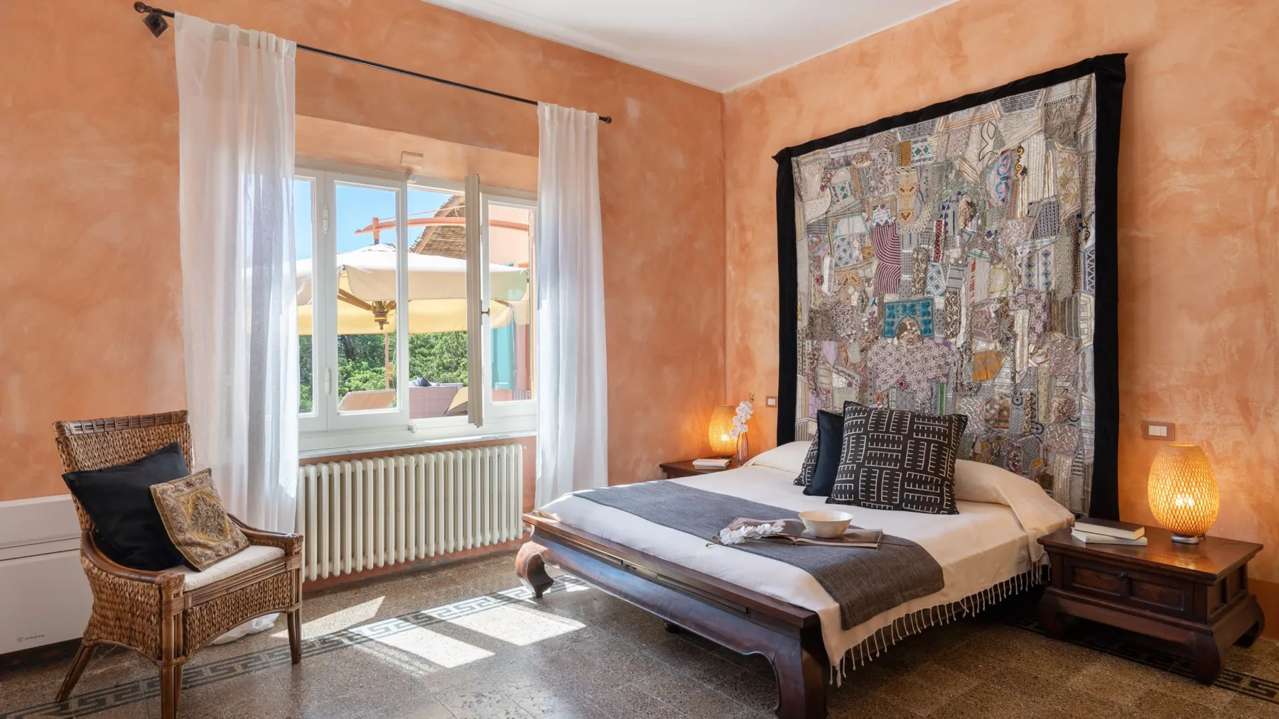 A bedroom with a bed, patterned wall hanging, and window with sheer curtains.