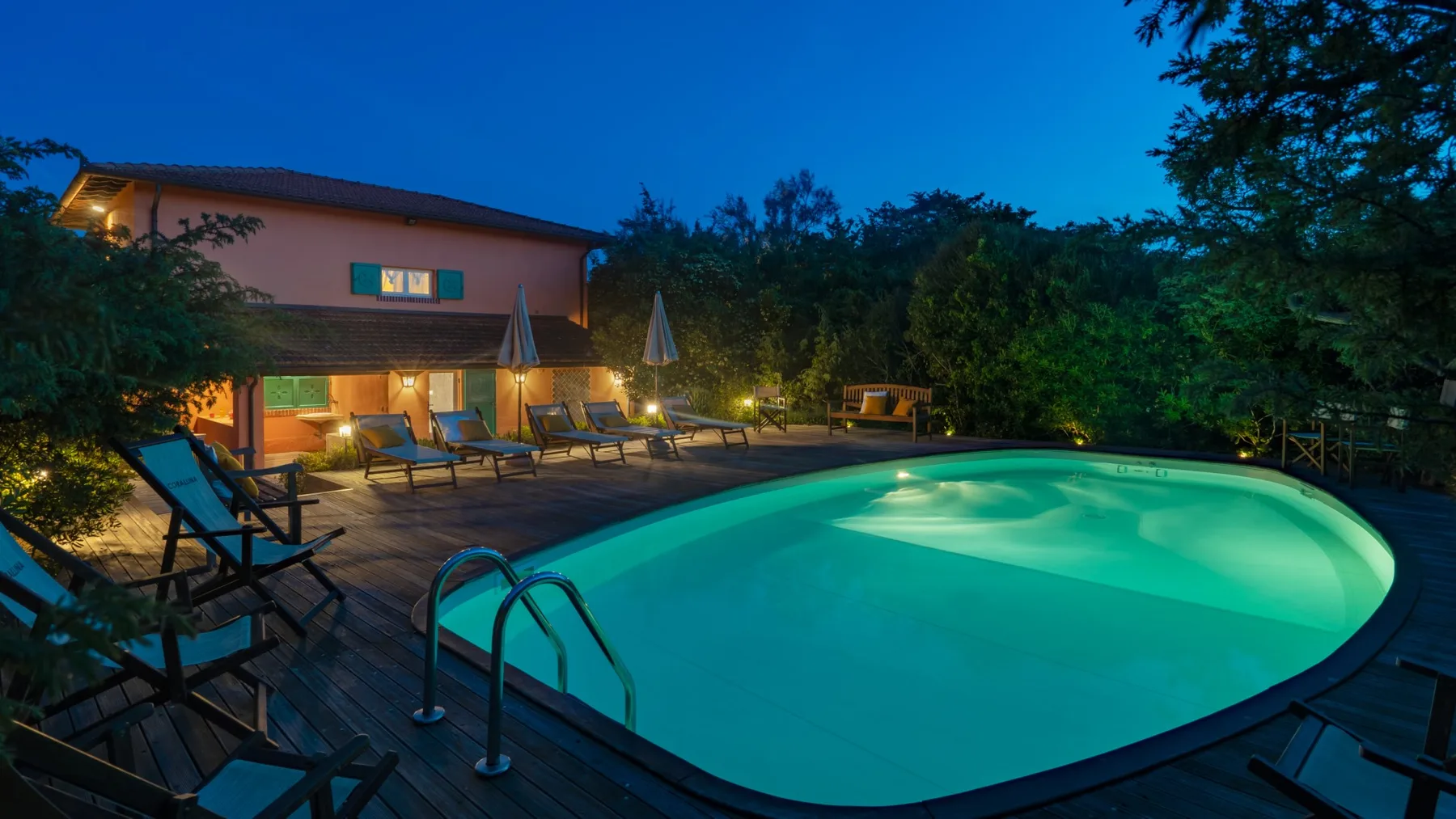 An illuminated swimming pool sits beside a wooden deck with lounge seating at dusk.