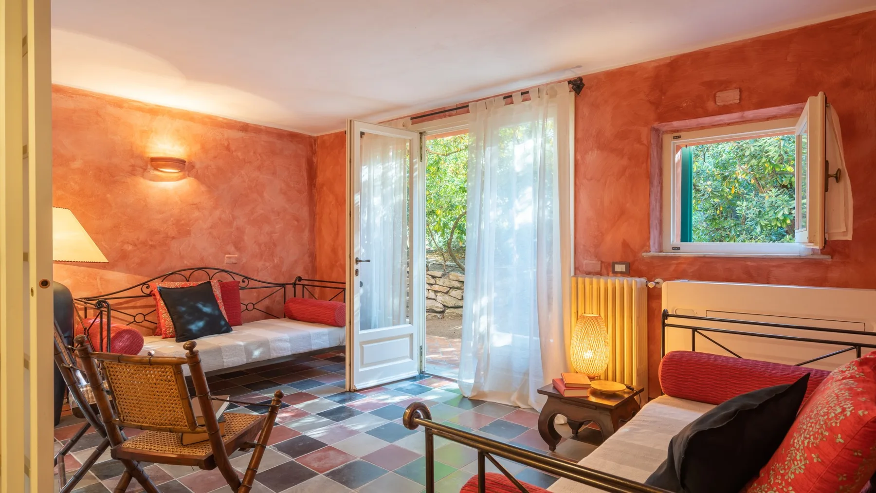 A warmly colored bedroom has two beds, seating, and open windows overlooking greenery.