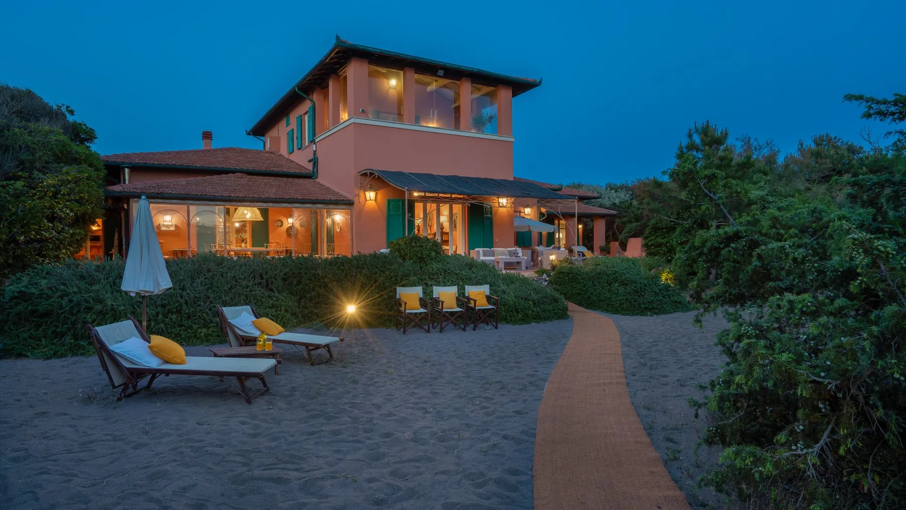 A modern house with a lit patio and a path through landscaped grounds at dusk.
