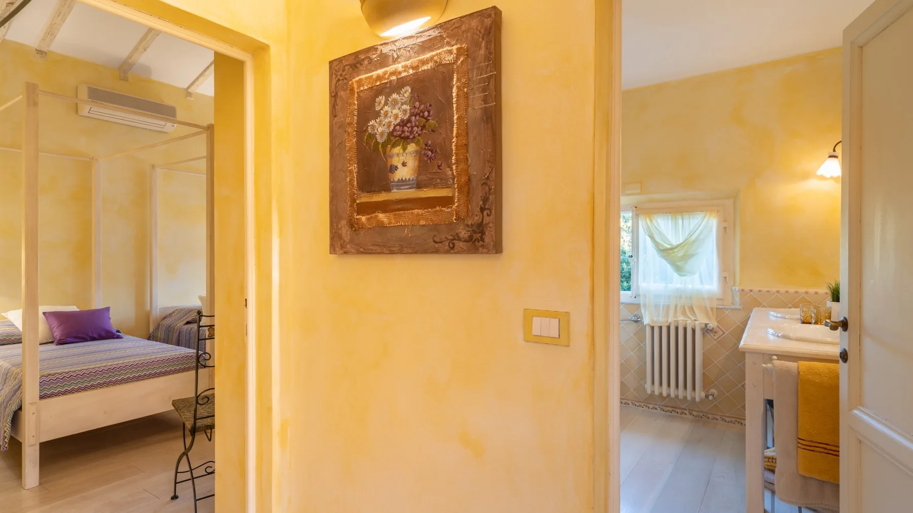 A warm-toned hallway connects a bedroom and bathroom, with floral artwork on the wall.