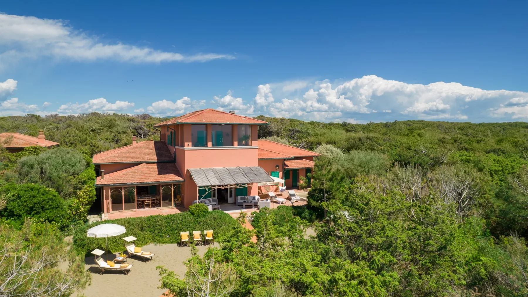 A two-story house with red-tiled roofs sits among lush greenery under a blue sky.