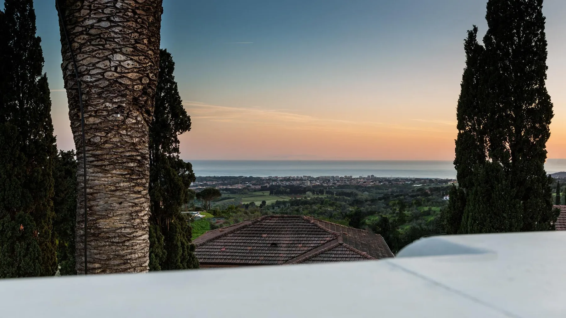 A sunset view over a tiled roof, trees, and the distant coastline.