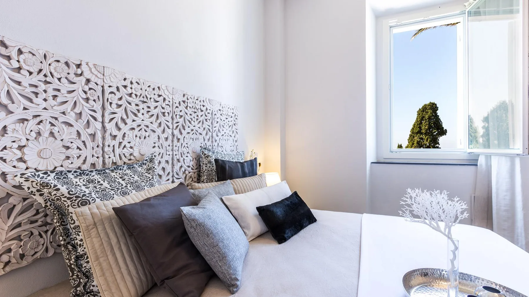 A bedroom with a bed, patterned cushions, and a bright window.