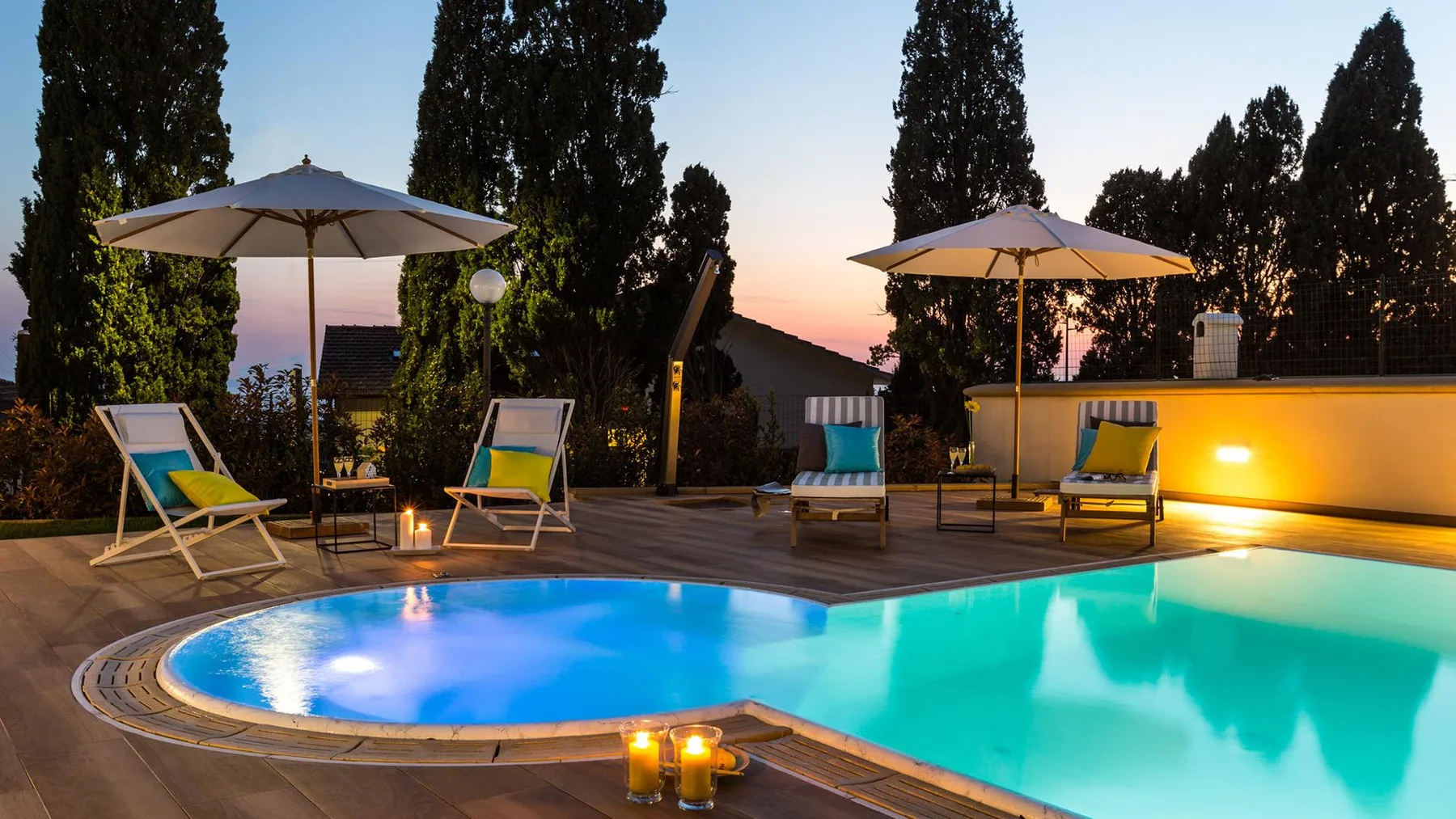 An outdoor swimming pool glows at dusk beside lounge seating and umbrellas.