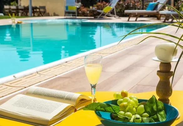 A swimming pool beside a sunlit patio, with a book and fruit in the foreground.