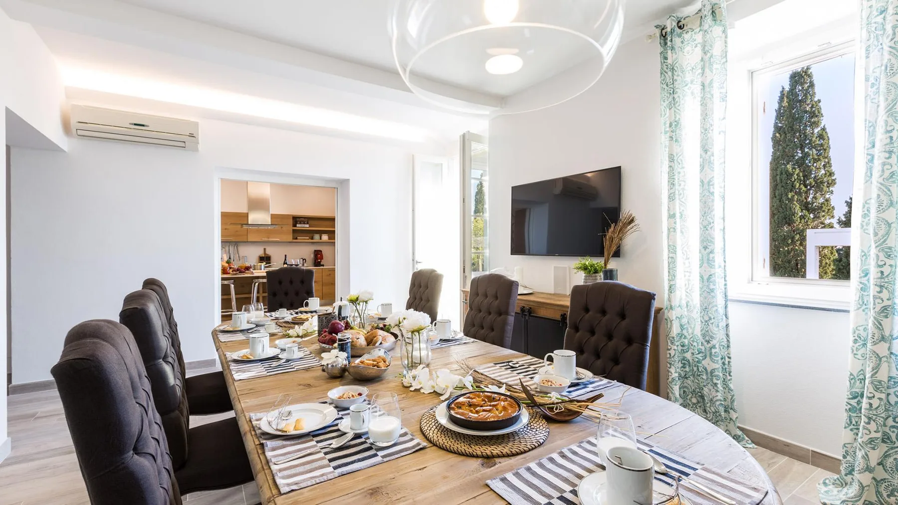 A dining area with a long table set for a meal beside large windows.