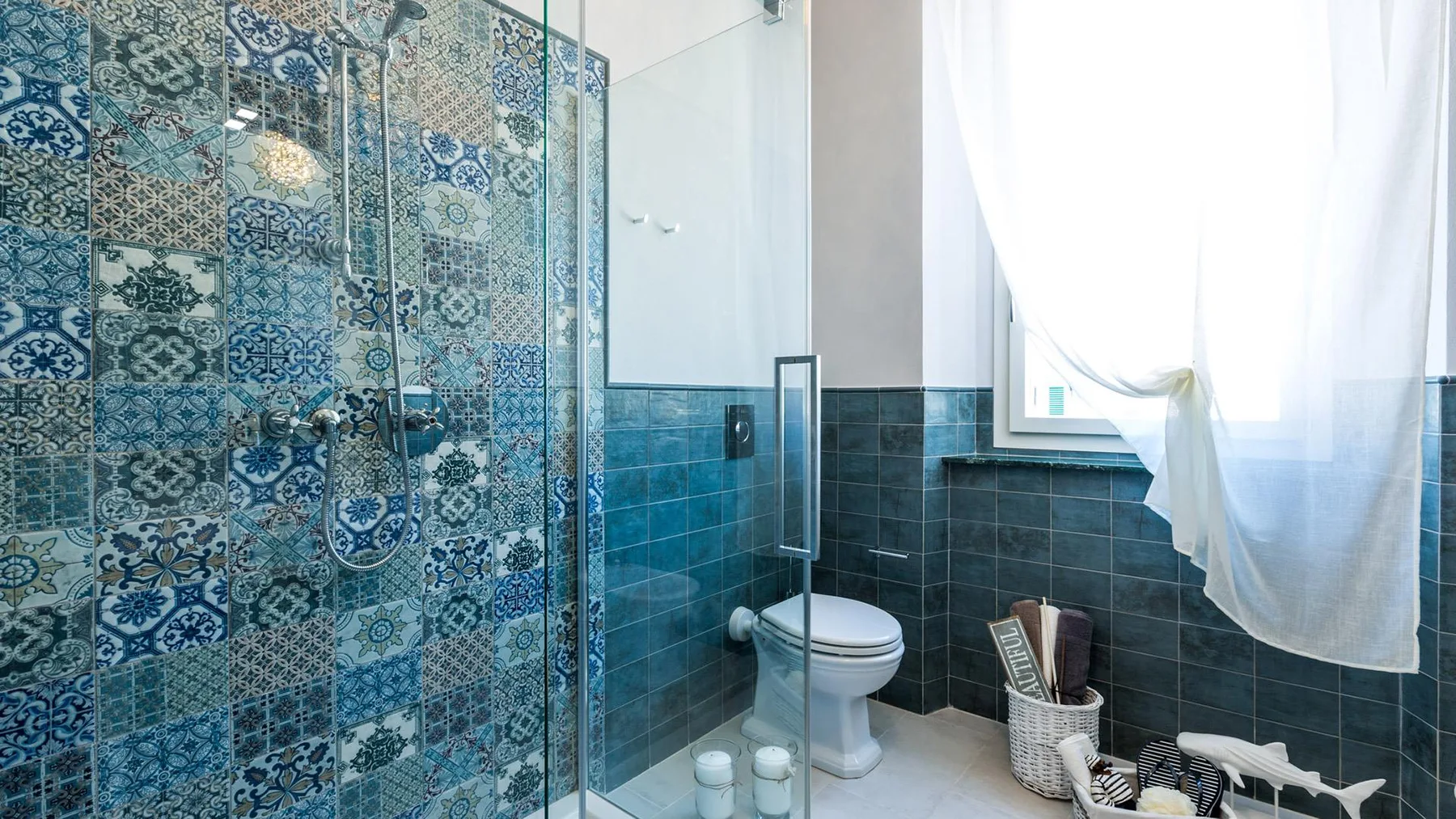 A bathroom with a glass shower, patterned tile, and a toilet beside a window.