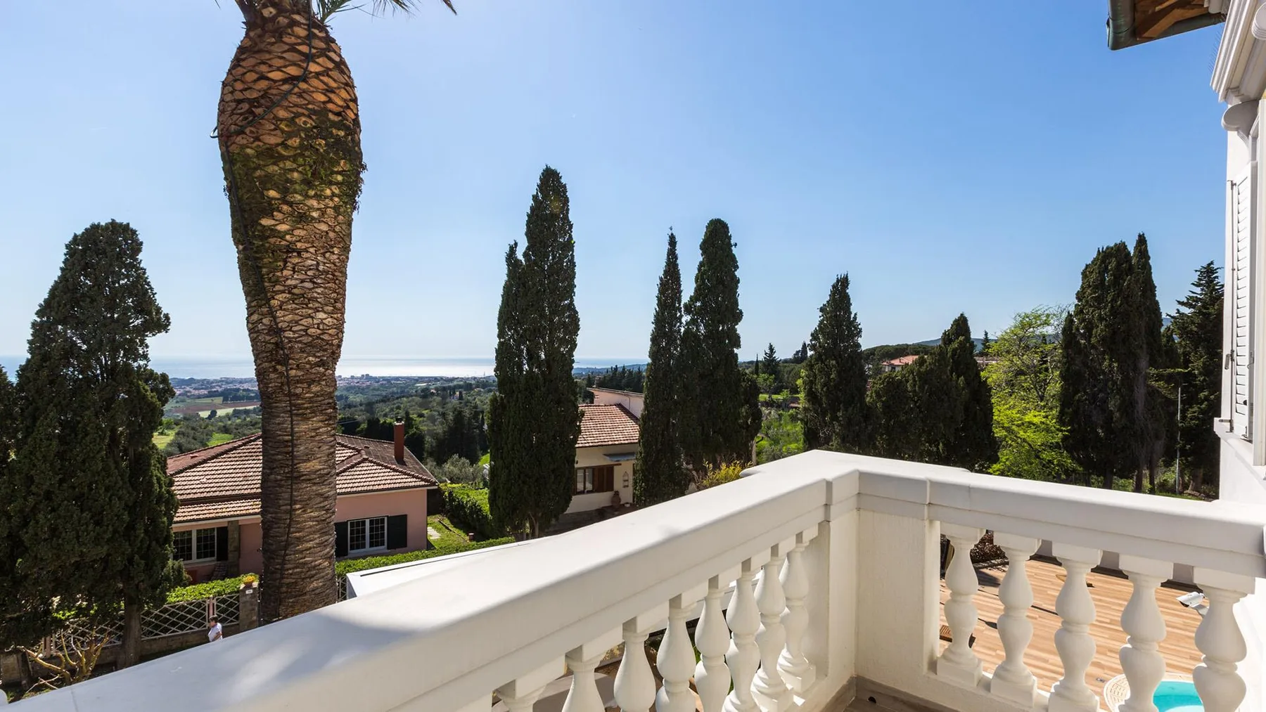 A white balcony railing overlooks nearby homes, trees, and a distant coastline.