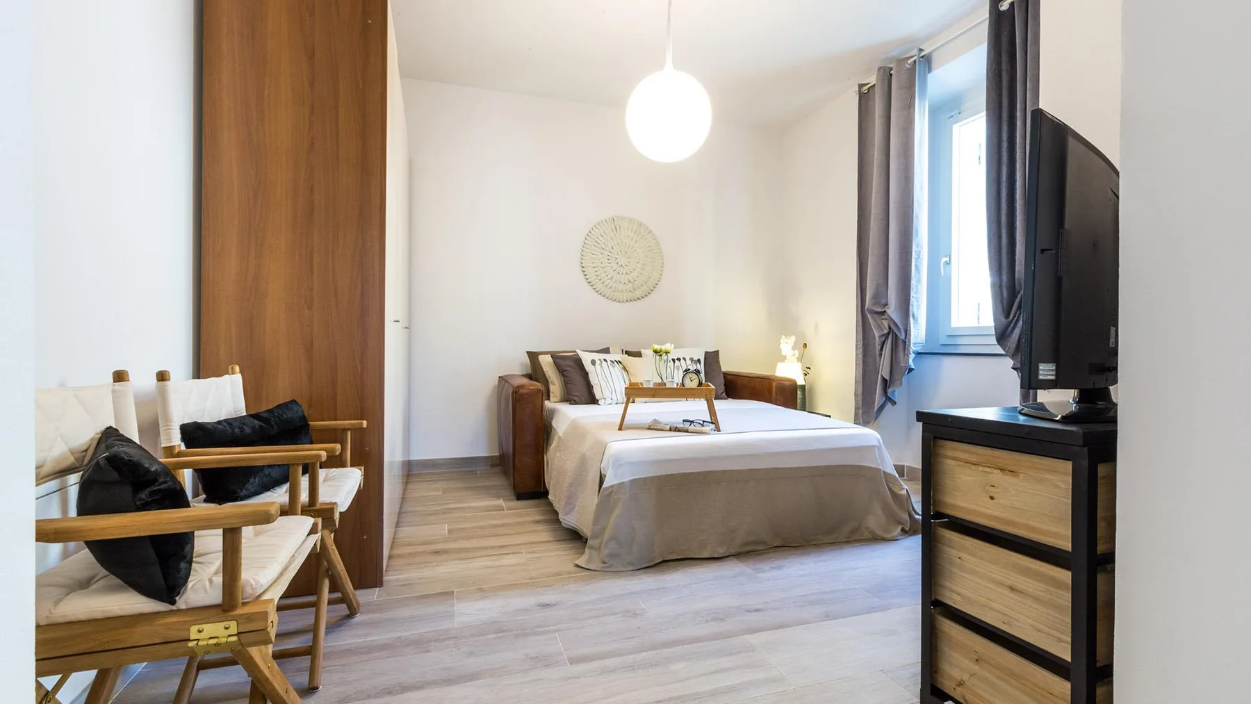 A bright bedroom with a bed, seating, and a television beside the window.