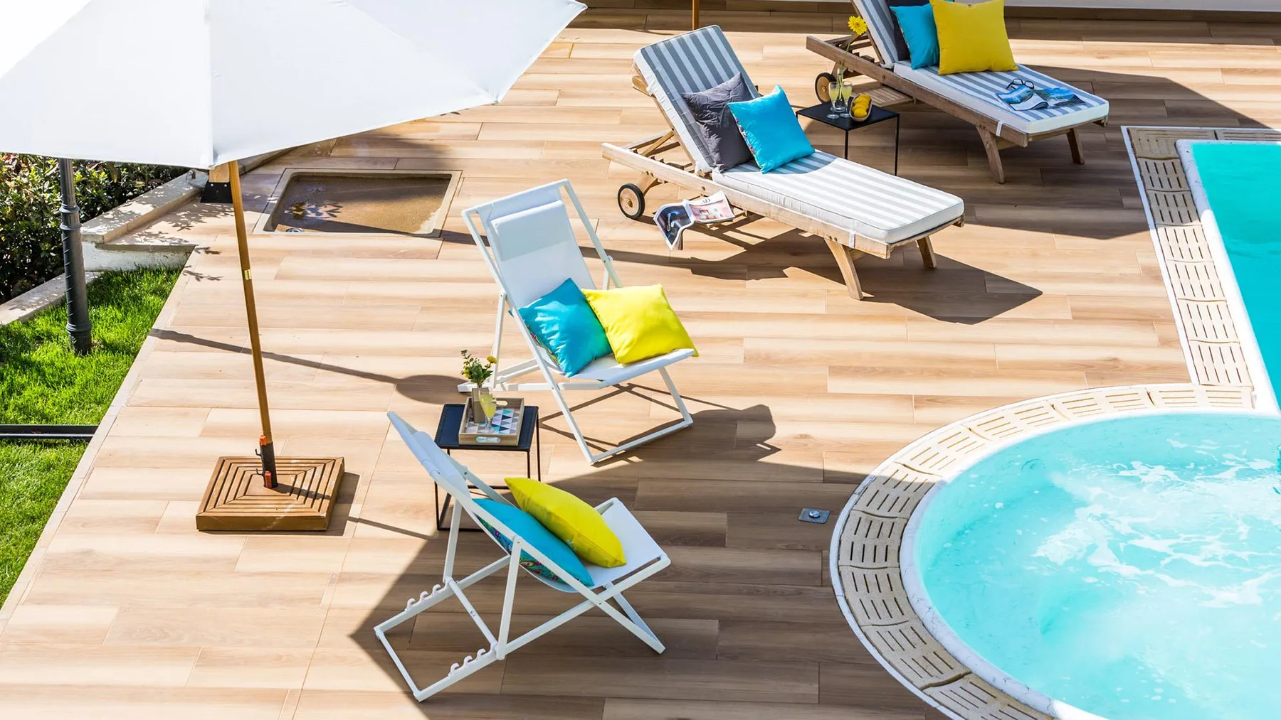 A sunny poolside deck has lounge chairs, colorful cushions, and a large white umbrella.