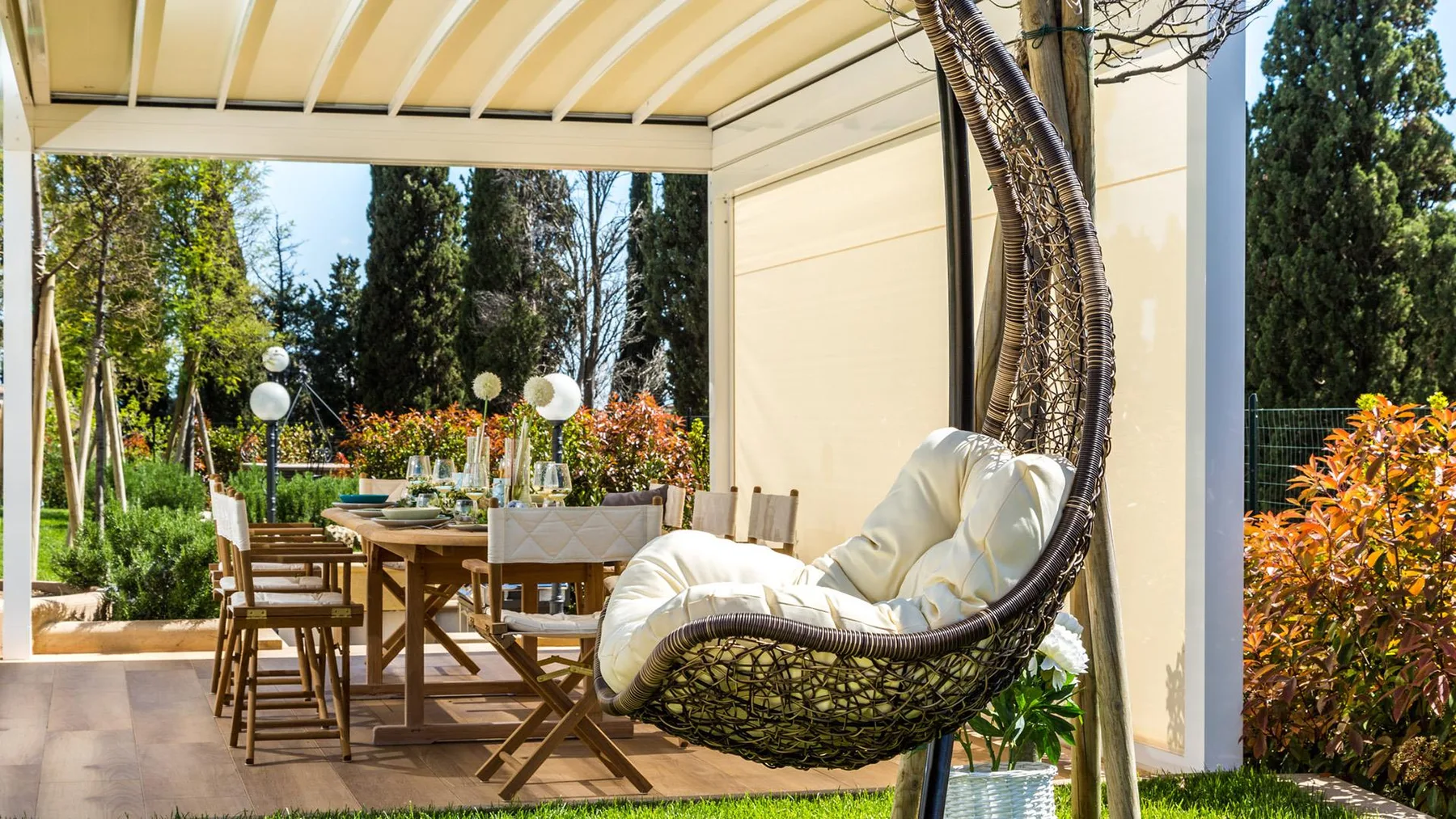 A covered outdoor dining area with a hanging woven seat and garden views.