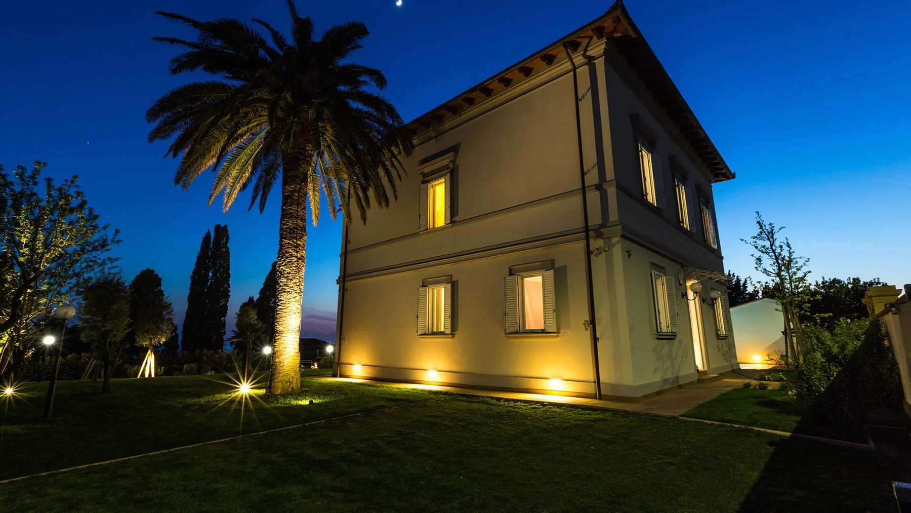 A two-story house at night, with lit windows and a palm tree in the garden.