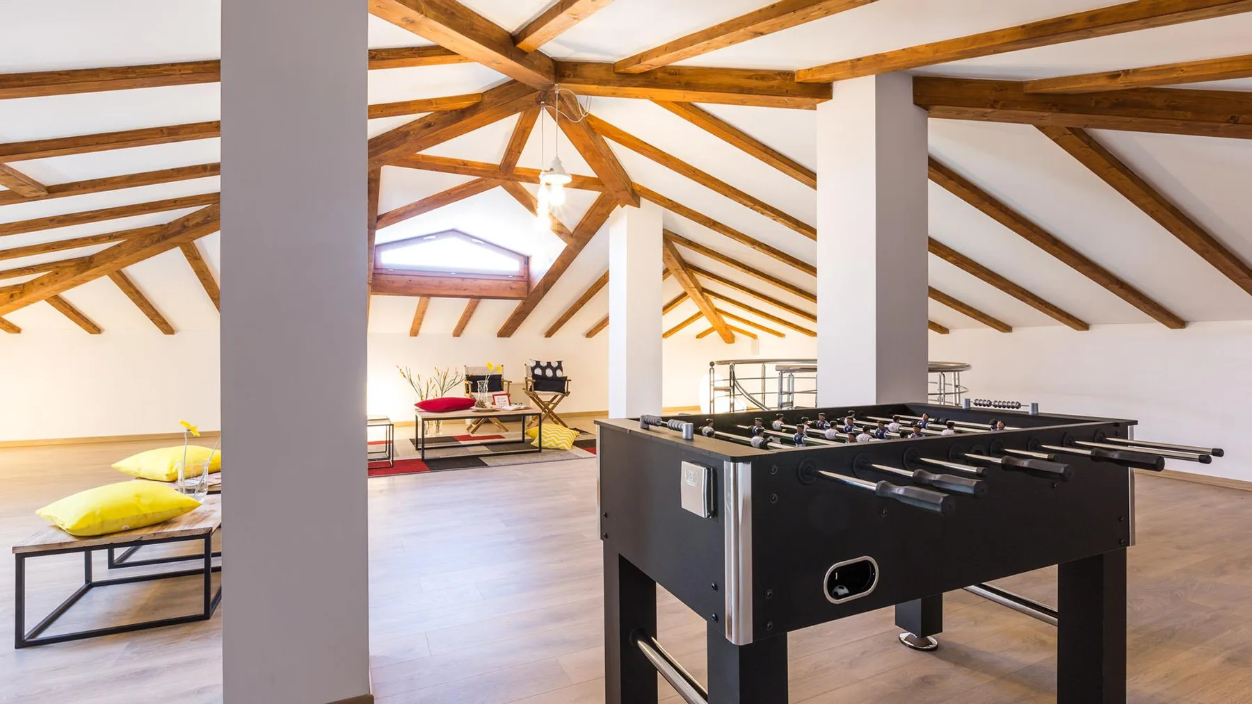 A bright loft game room has a foosball table beneath exposed wooden ceiling beams.