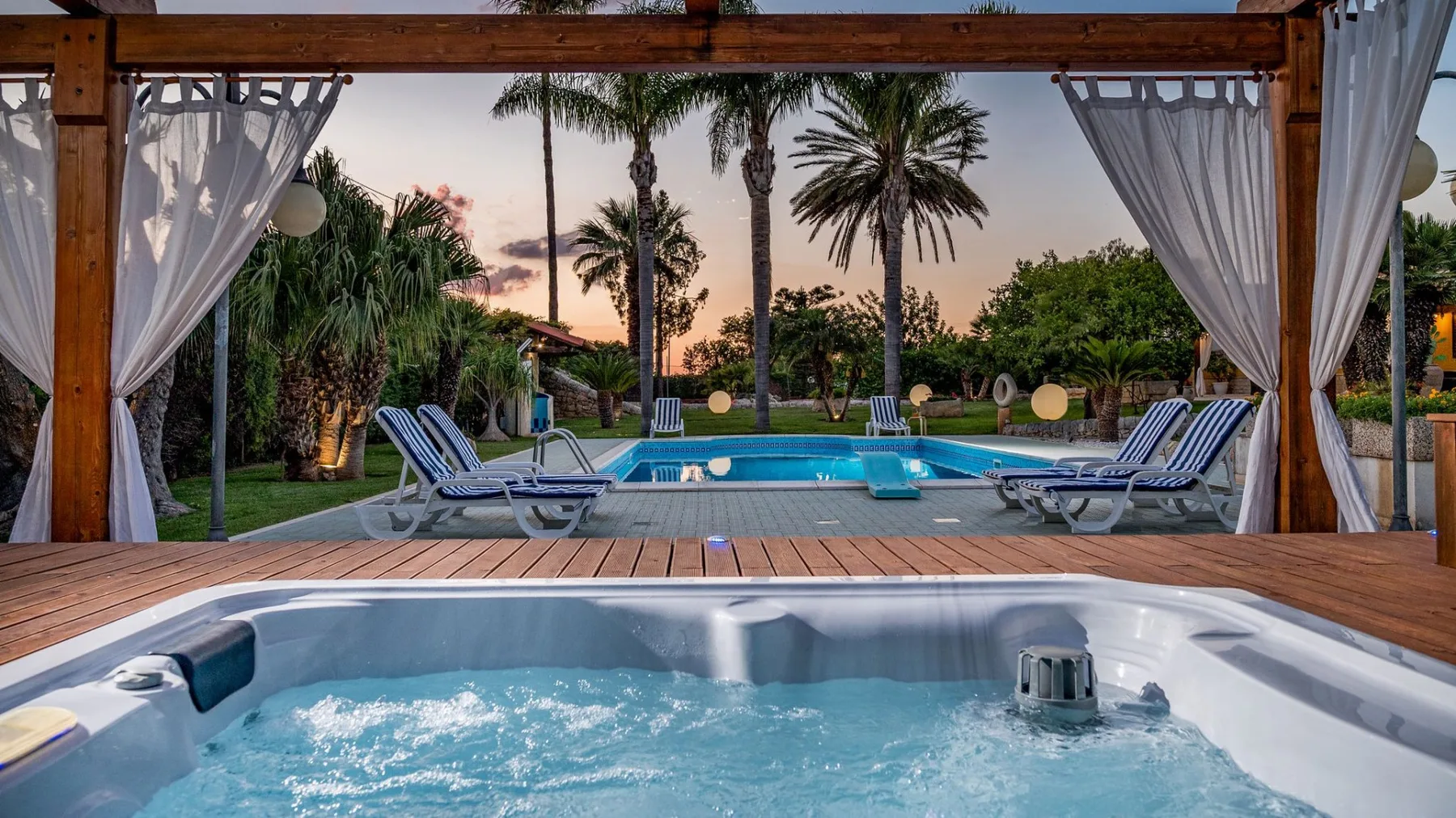 Una piscina y un jacuzzi burbujeante están junto a un patio de madera cubierto al anochecer.
