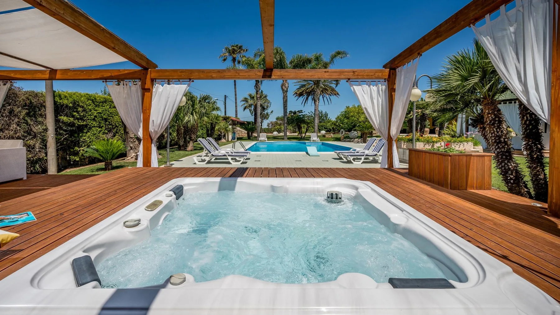 Un spa al aire libre está bajo una pérgola de madera, con una piscina al fondo.