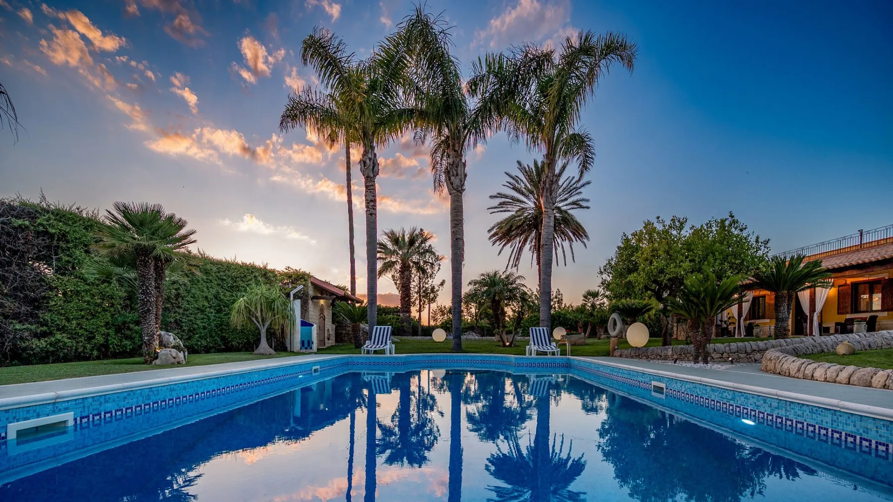 Una piscina al aire libre refleja palmeras bajo un cielo colorido al atardecer.
