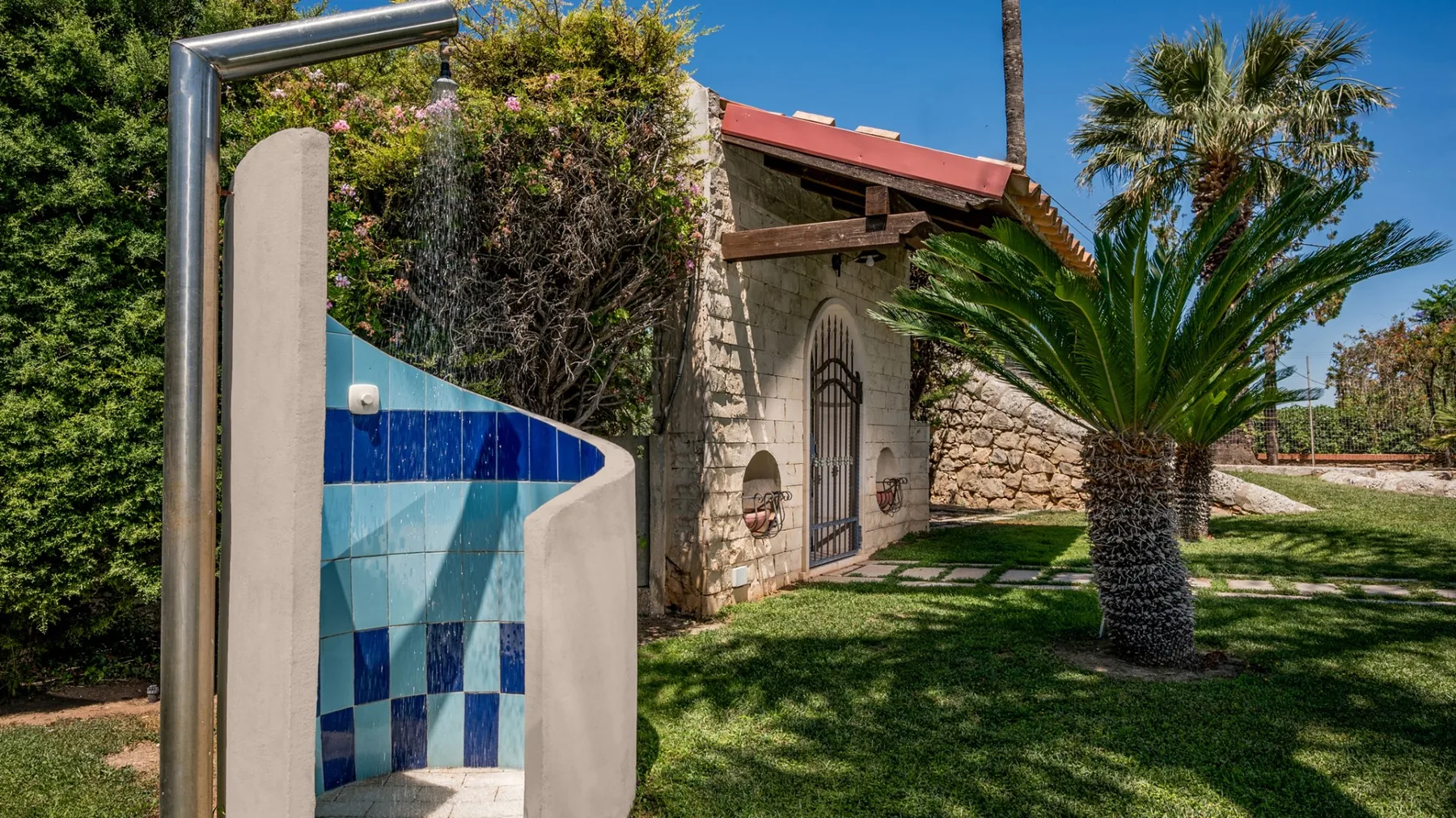 Una construcción de piedra está junto a una ducha exterior con azulejos y una palmera.