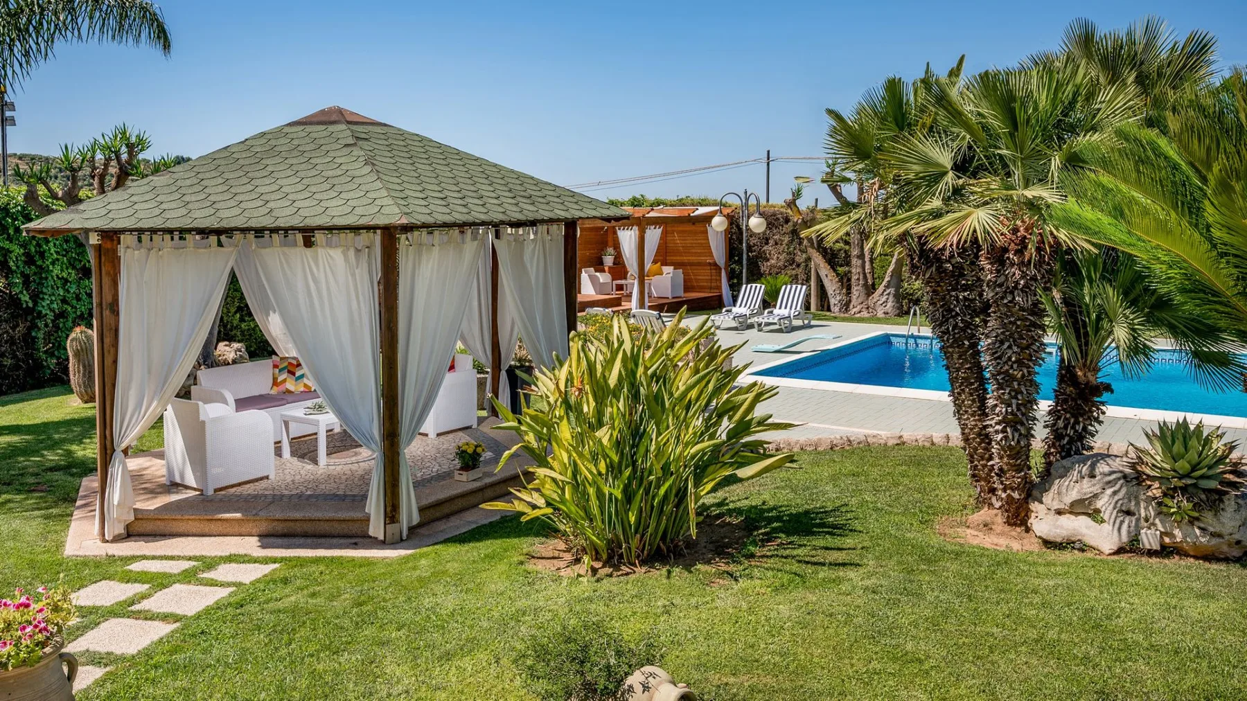 Zona de piscina ajardinada con una cabaña sombreada, tumbonas y palmeras.