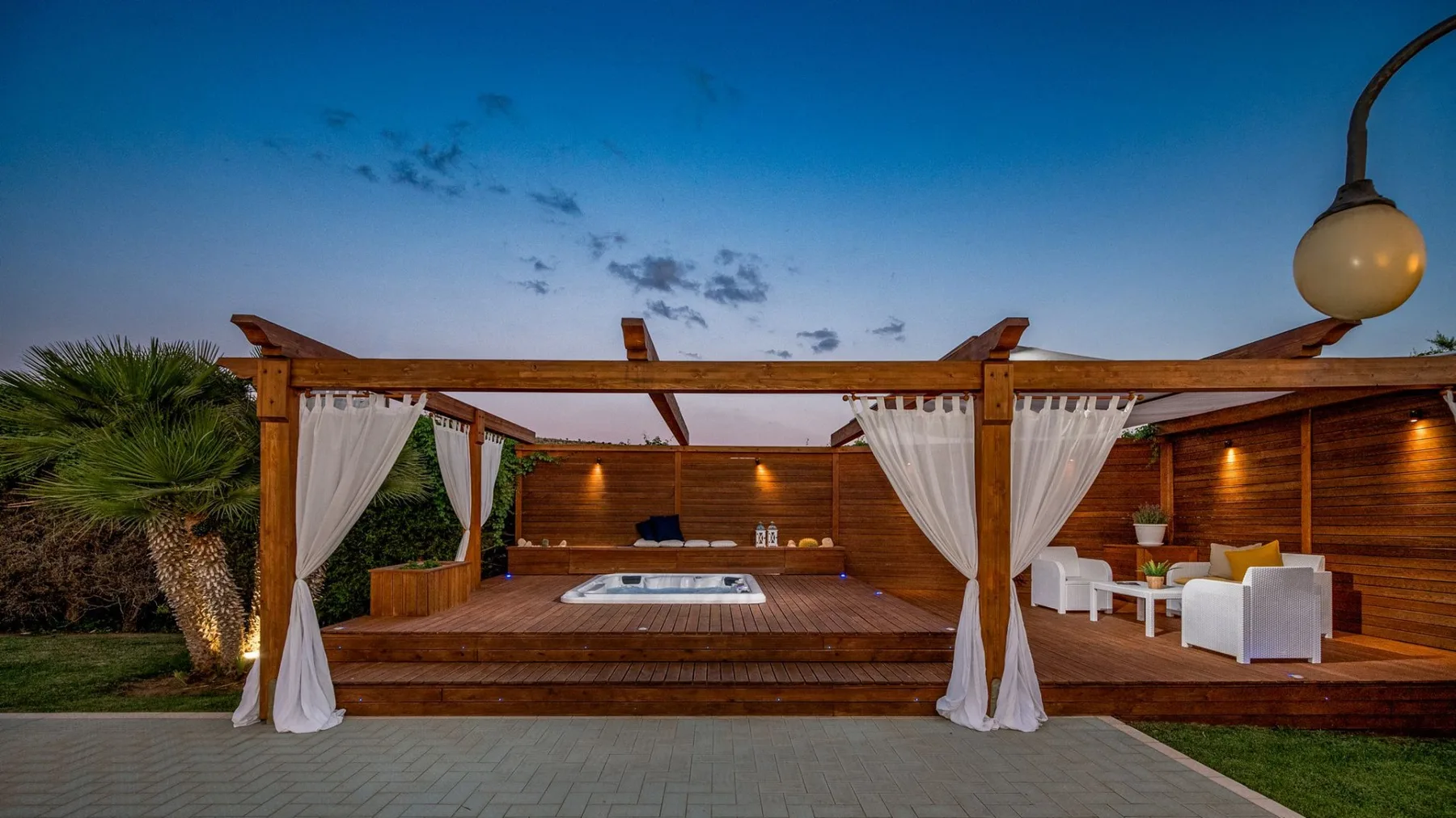 Patio exterior con bañera de hidromasaje, asientos y pérgola de madera con cortinas al anochecer.