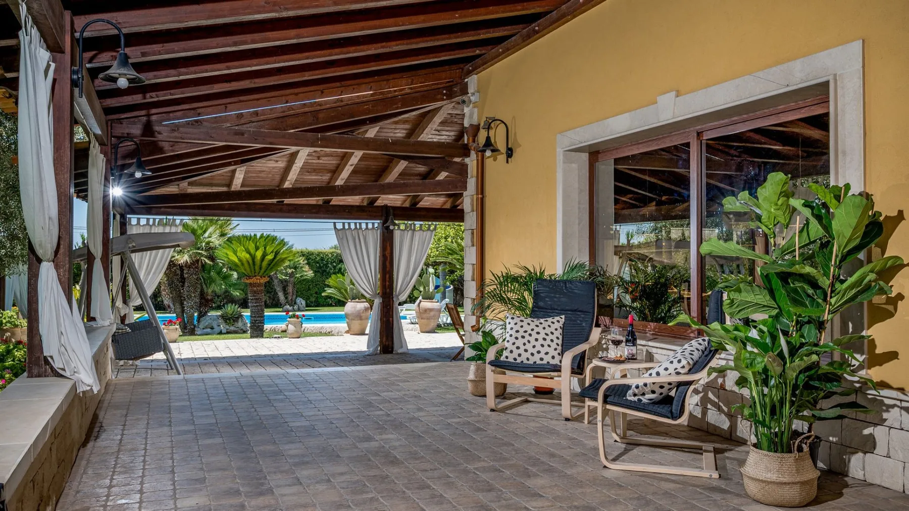 Un patio cubierto con suelo de baldosas, asientos y vistas a una piscina y un jardín.