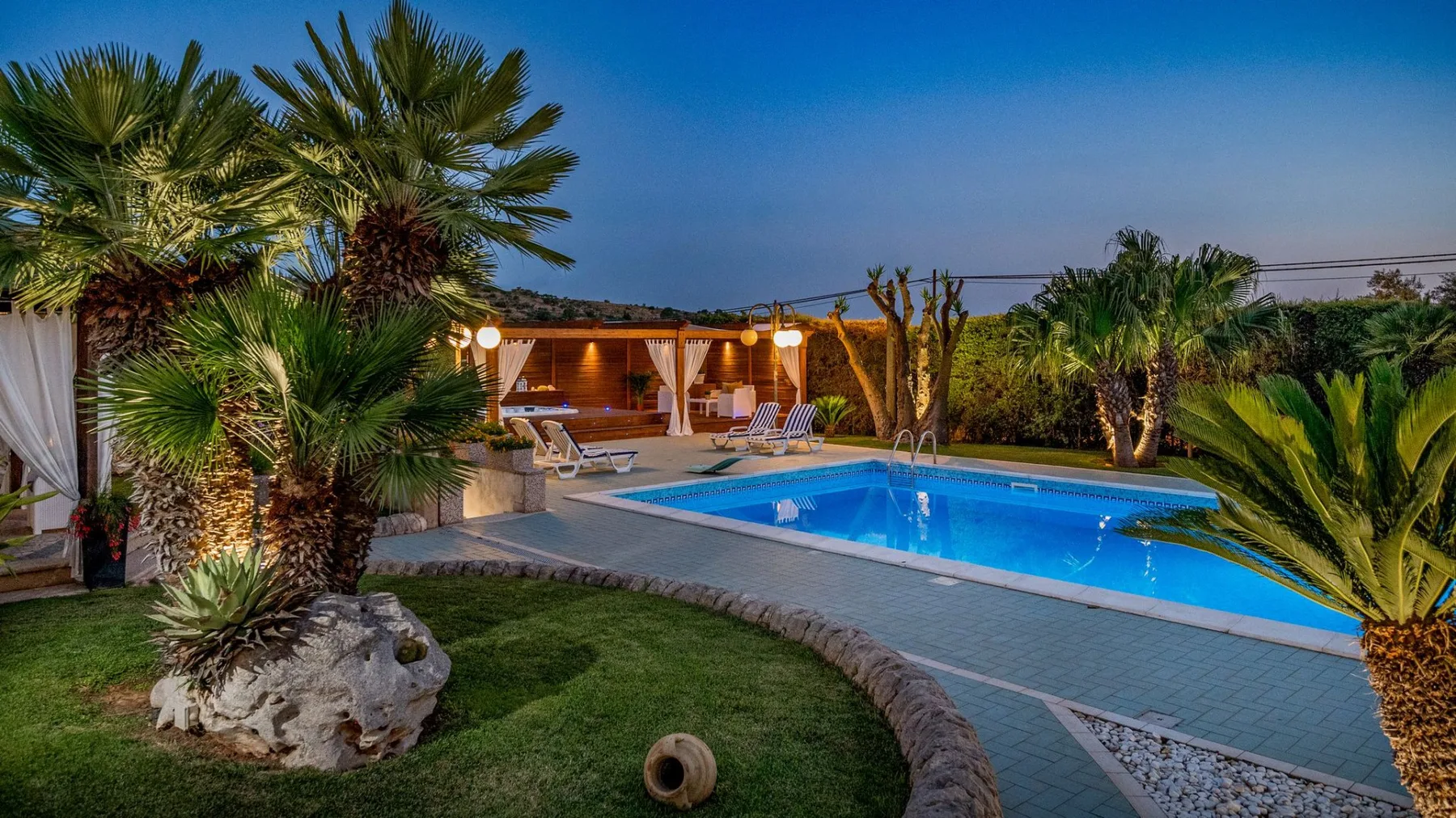 Una piscina al aire libre está rodeada de césped, palmeras y asientos.