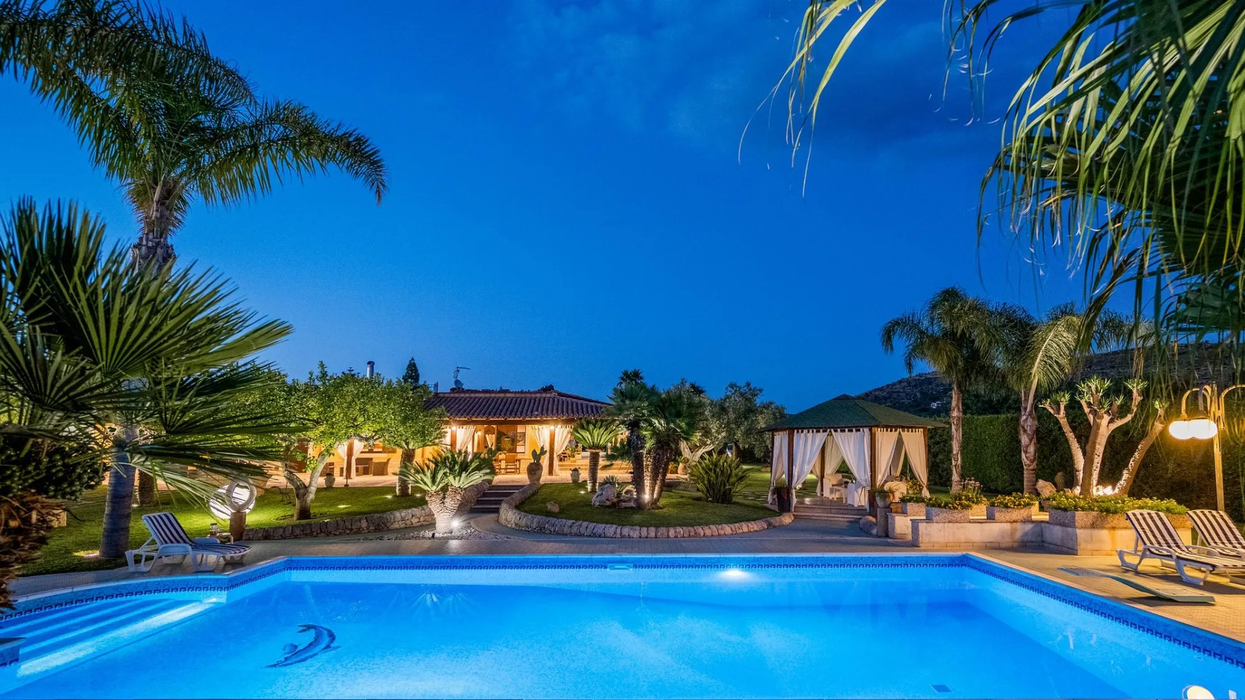 Una piscina está en un jardín tropical junto a una zona de estar exterior iluminada.