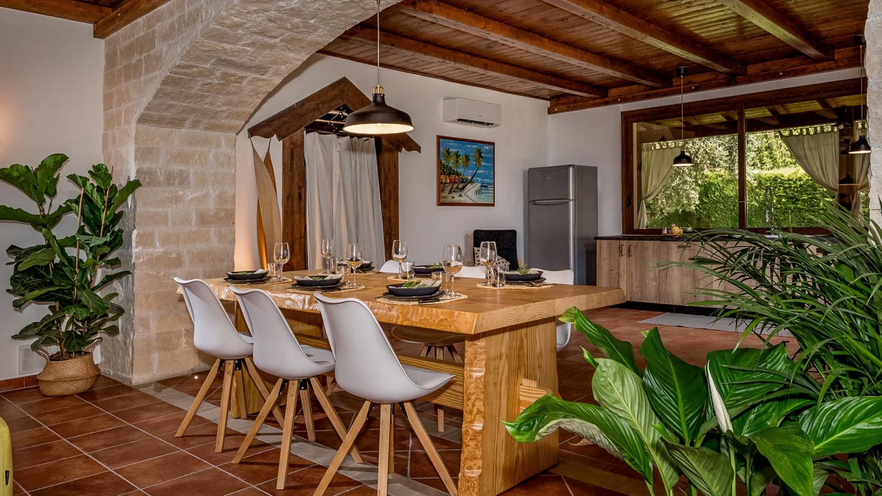 Un comedor con una mesa larga de madera, asientos blancos y plantas frondosas.