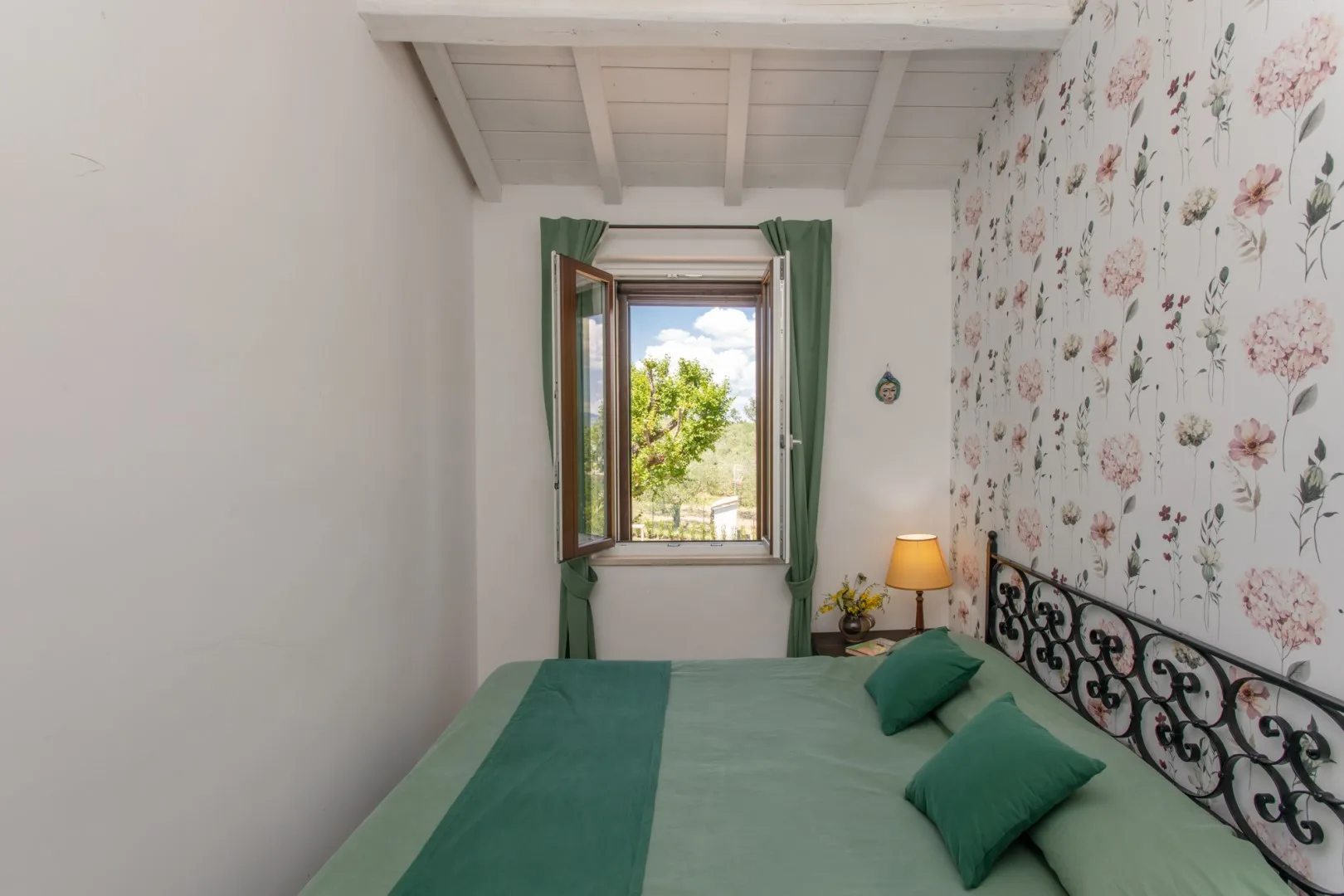 A bedroom with a green-covered bed, floral wallpaper, and an open window.