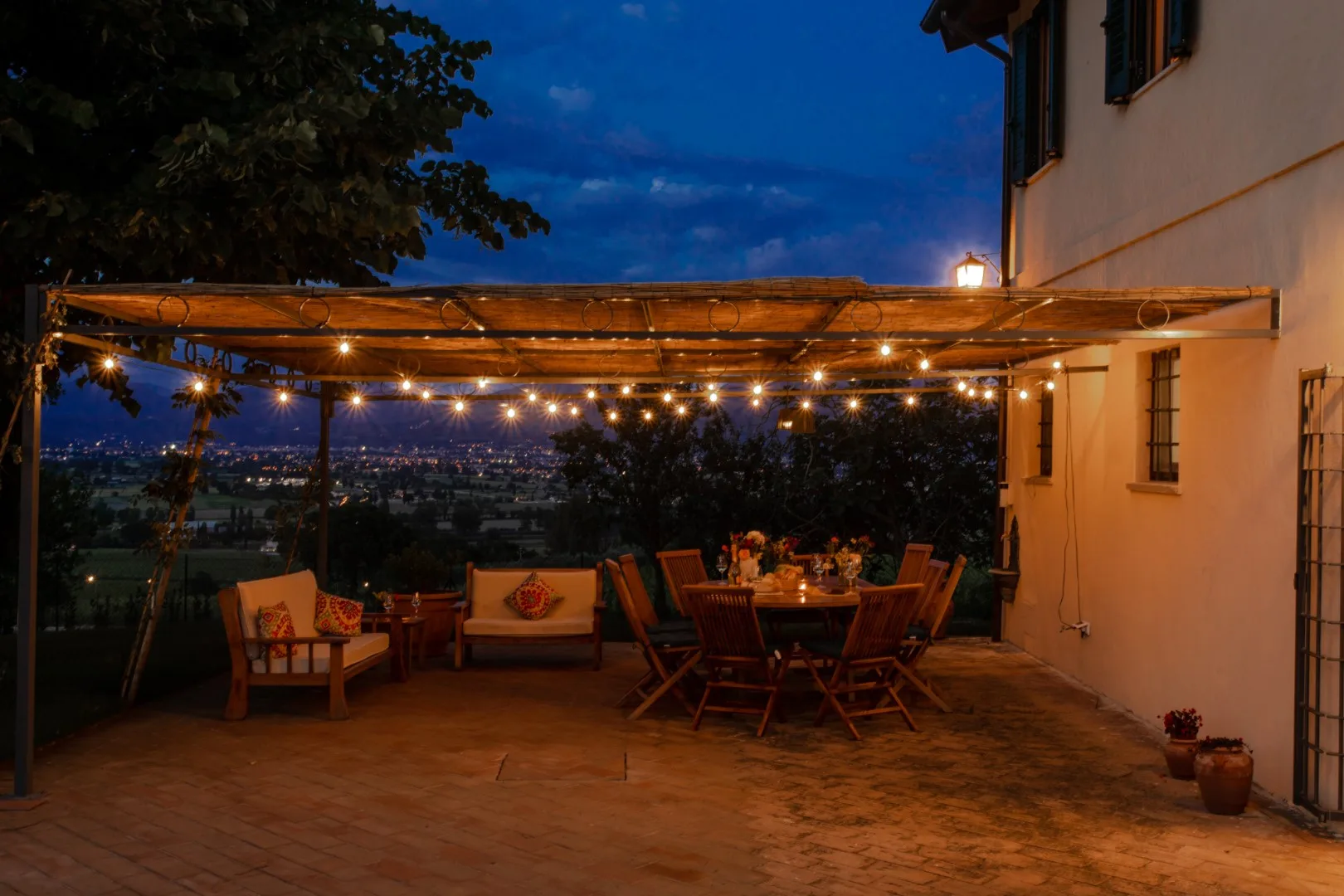 A warmly lit outdoor dining terrace beneath a pergola overlooks a city at dusk.