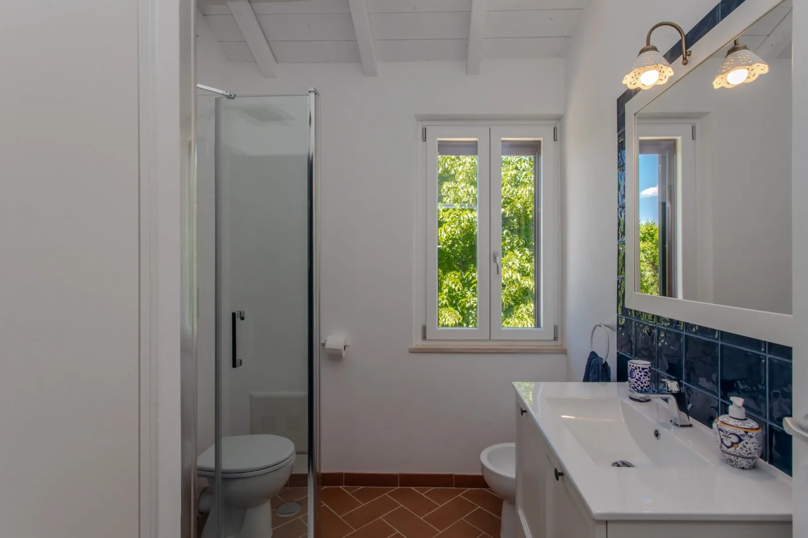 A bright bathroom with a glass shower, a double sink, and a window.