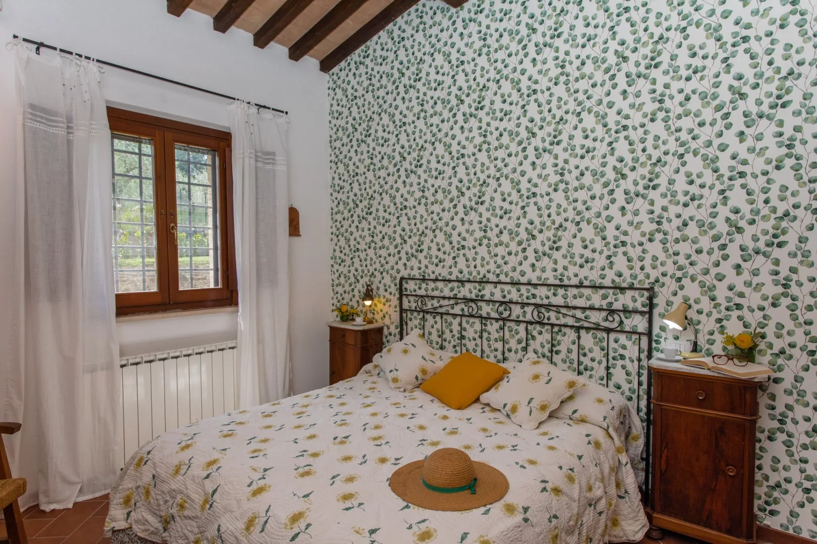 A bedroom with a patterned bedspread, a window, and leafy wallpaper.