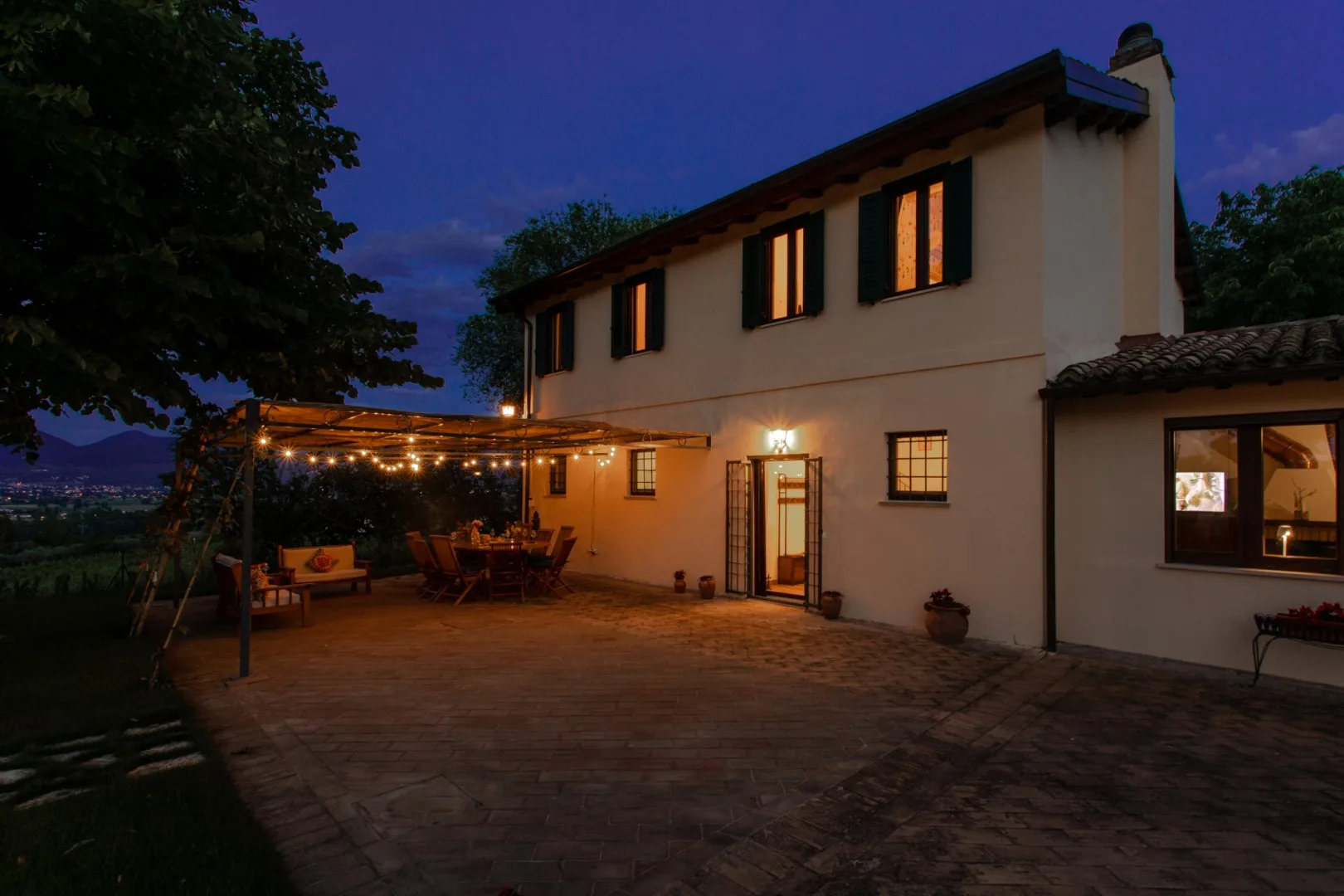 A two-story house opens onto a paved patio lit by warm string lights at dusk.